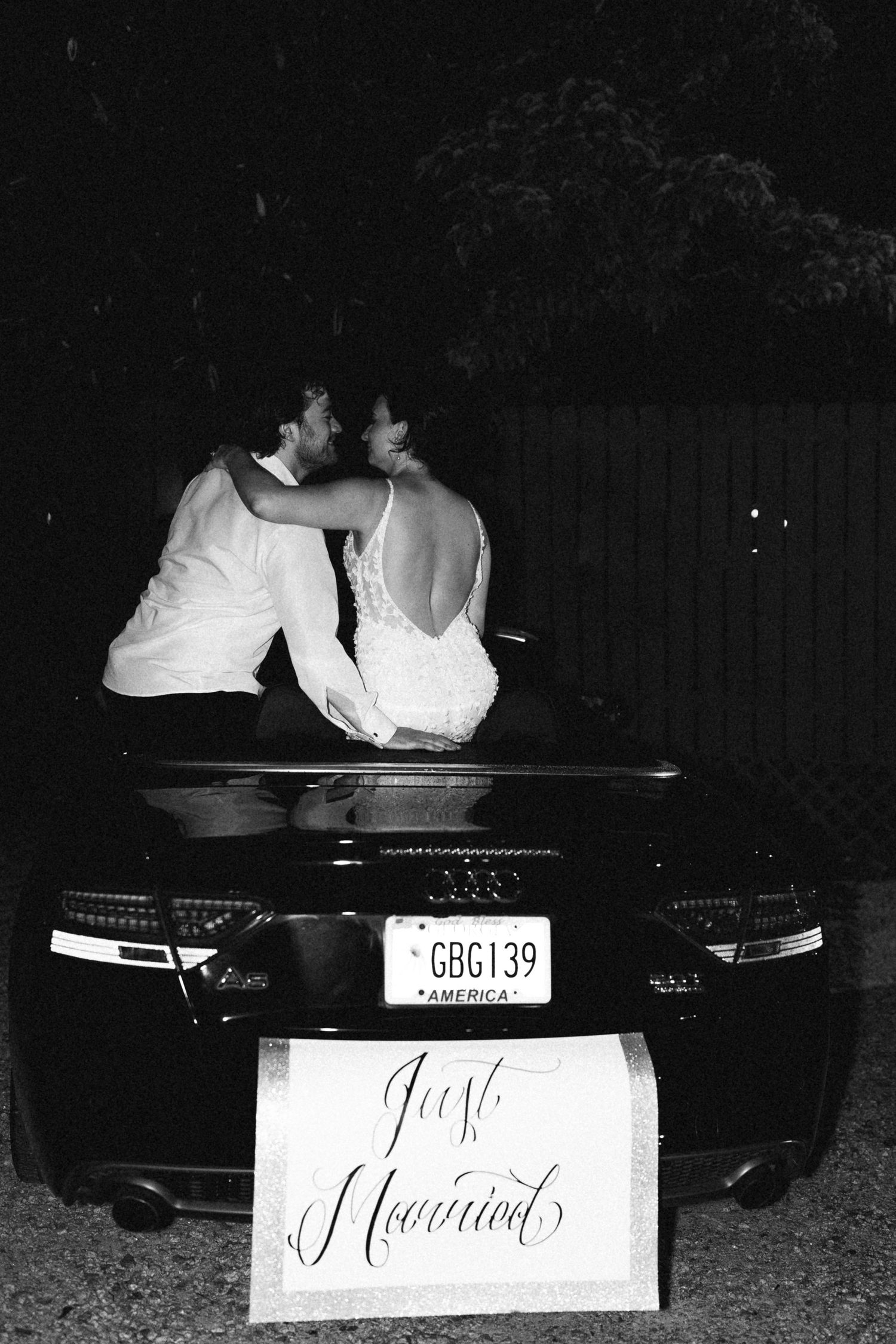 Black and white photo of newlyweds sitting on Audi car with Just Married sign at night.