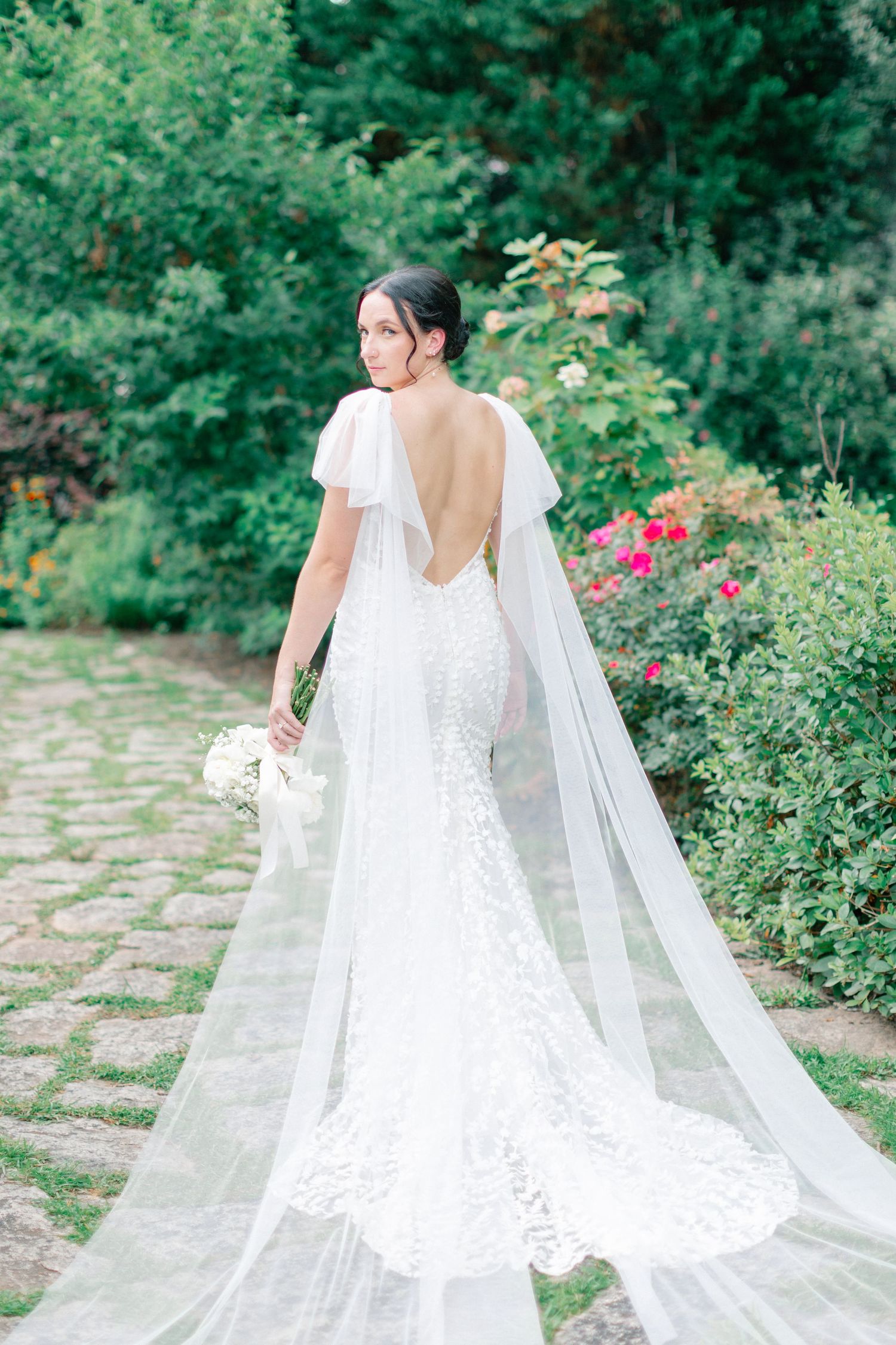 Bride in lace backless wedding gown with flowing veil holds bouquet in lush garden setting.