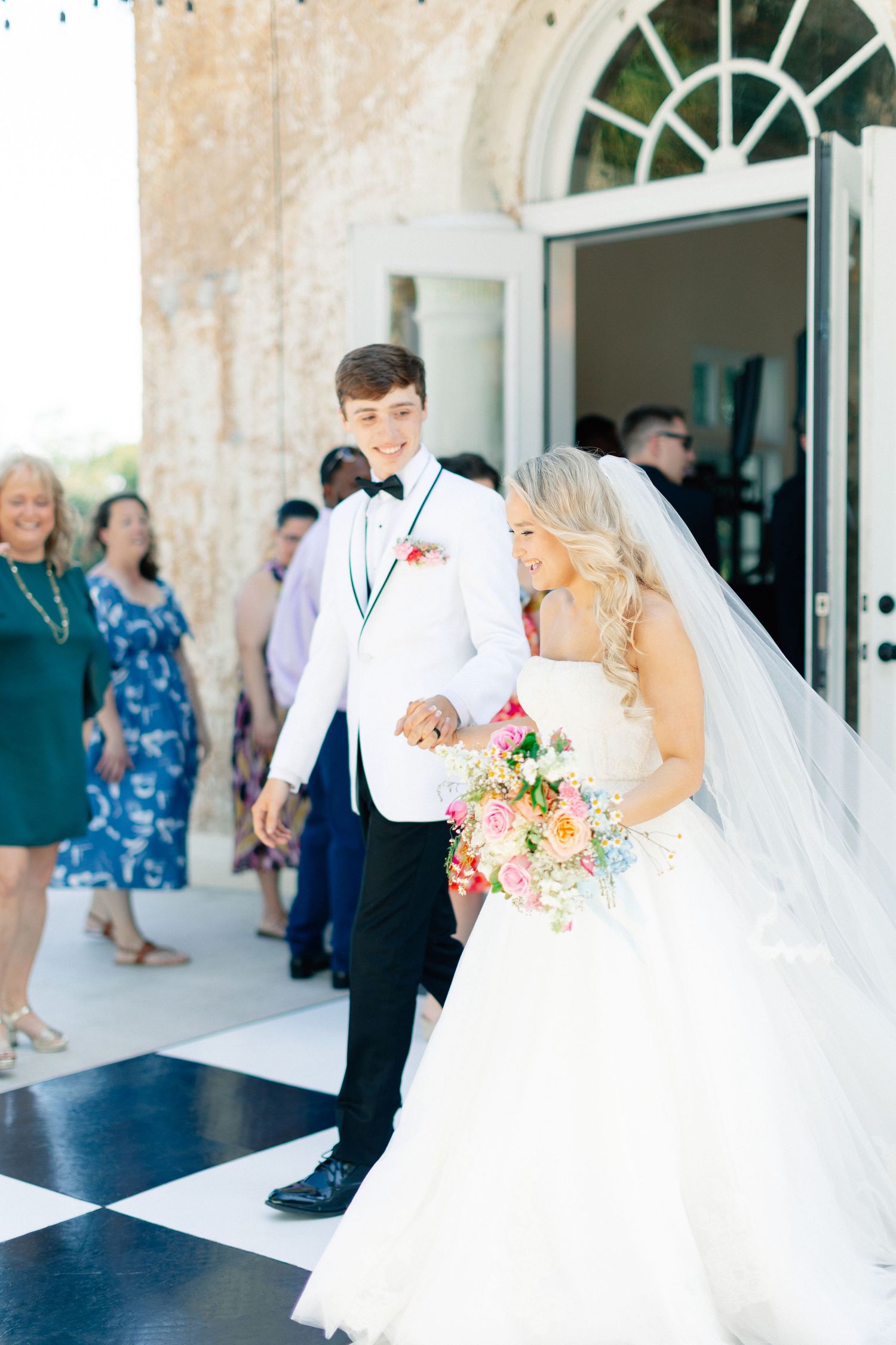 Bride and groom exit wedding venue smiling, bride holding colorful bouquet, groom in white tuxedo jacket, wedding at Bella Cosa