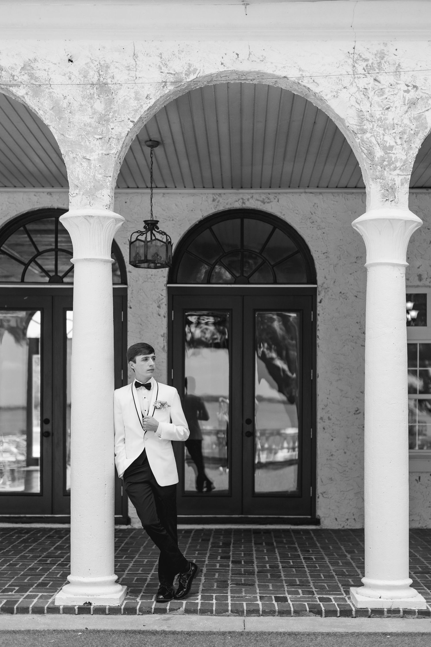 Elegant groom in white tuxedo jacket and bow tie leaning against column under arched doorway in black and white photo, wedding at Bella Cosa