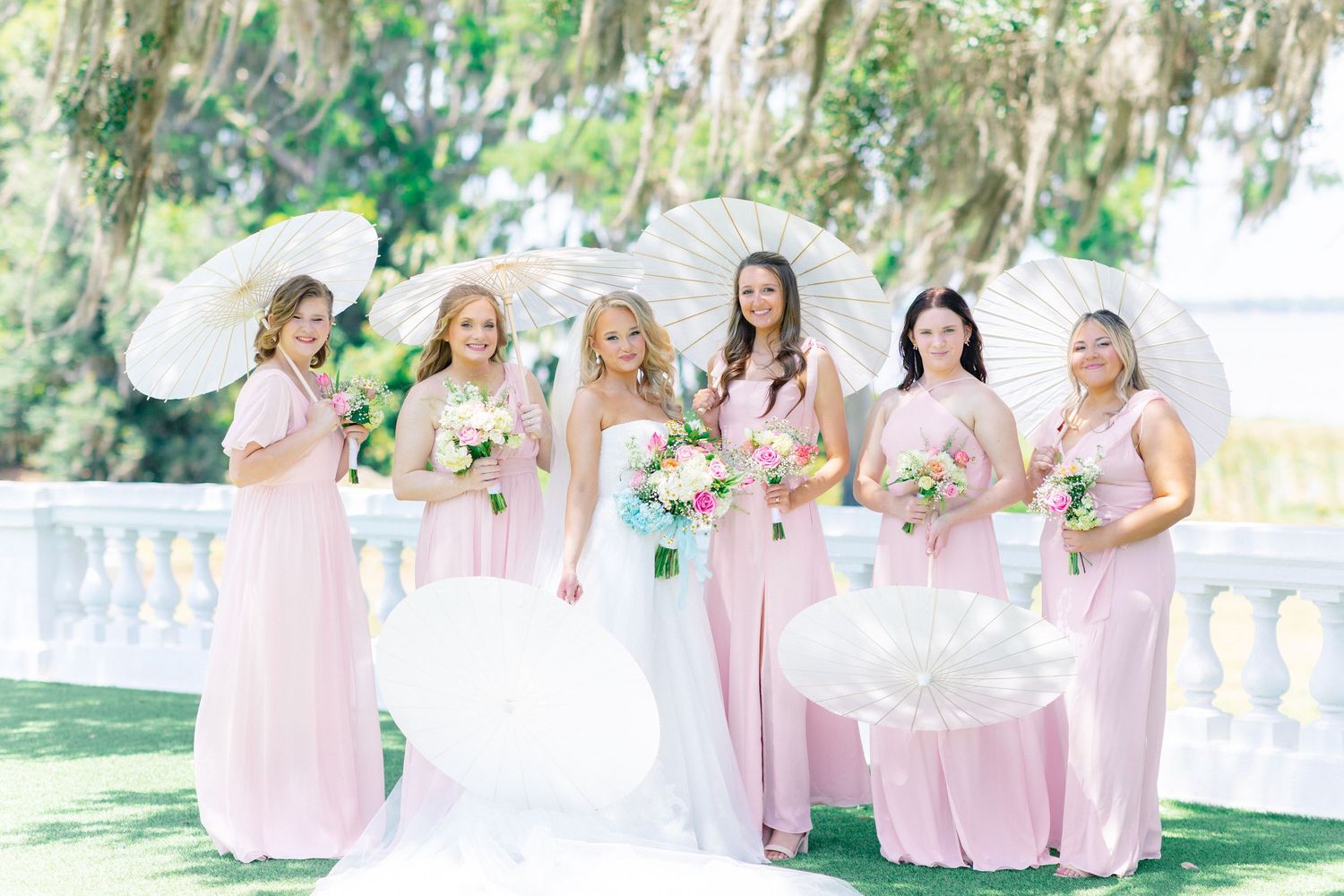 Bride in white gown with bridesmaids in pink dresses holding white parasols and floral bouquets outdoors, wedding at Bella Cosa