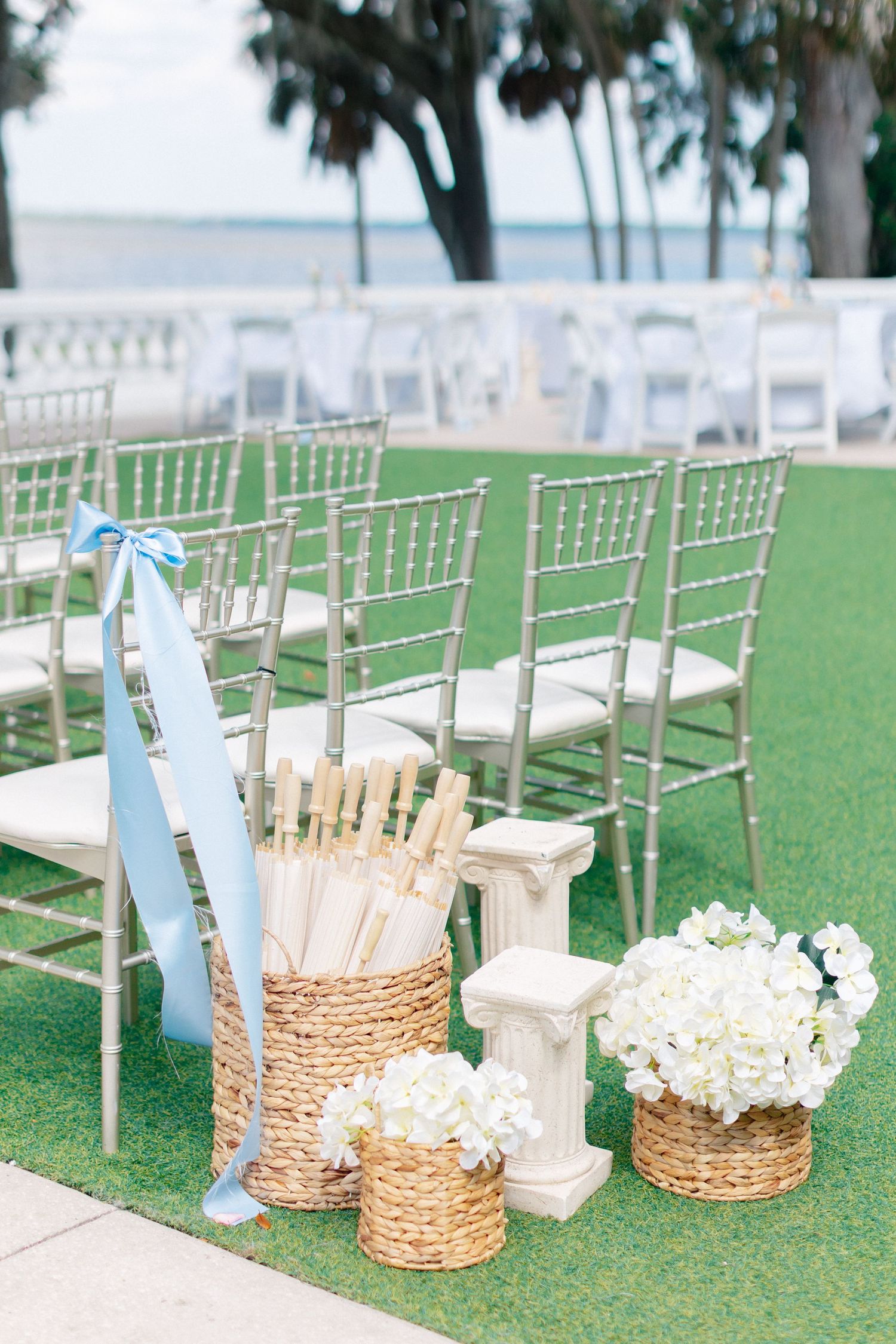 Outdoor wedding ceremony setup with silver chiavari chairs, wicker baskets of white flowers, and hand fans by the waterfront, Bella Cosa