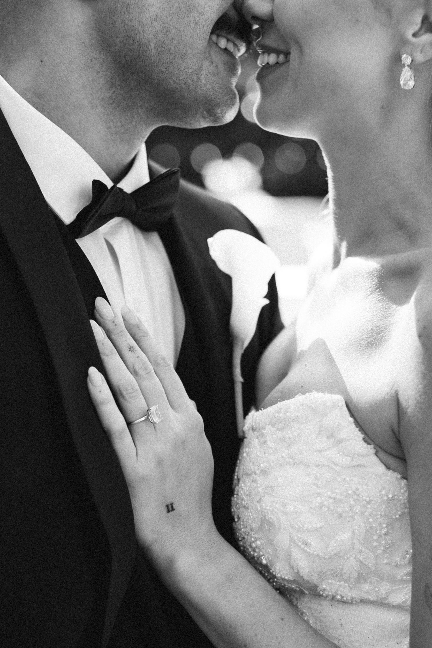 Black and white close-up of bride and groom nearly kissing, bride's hand showing diamond ring on groom's chest.