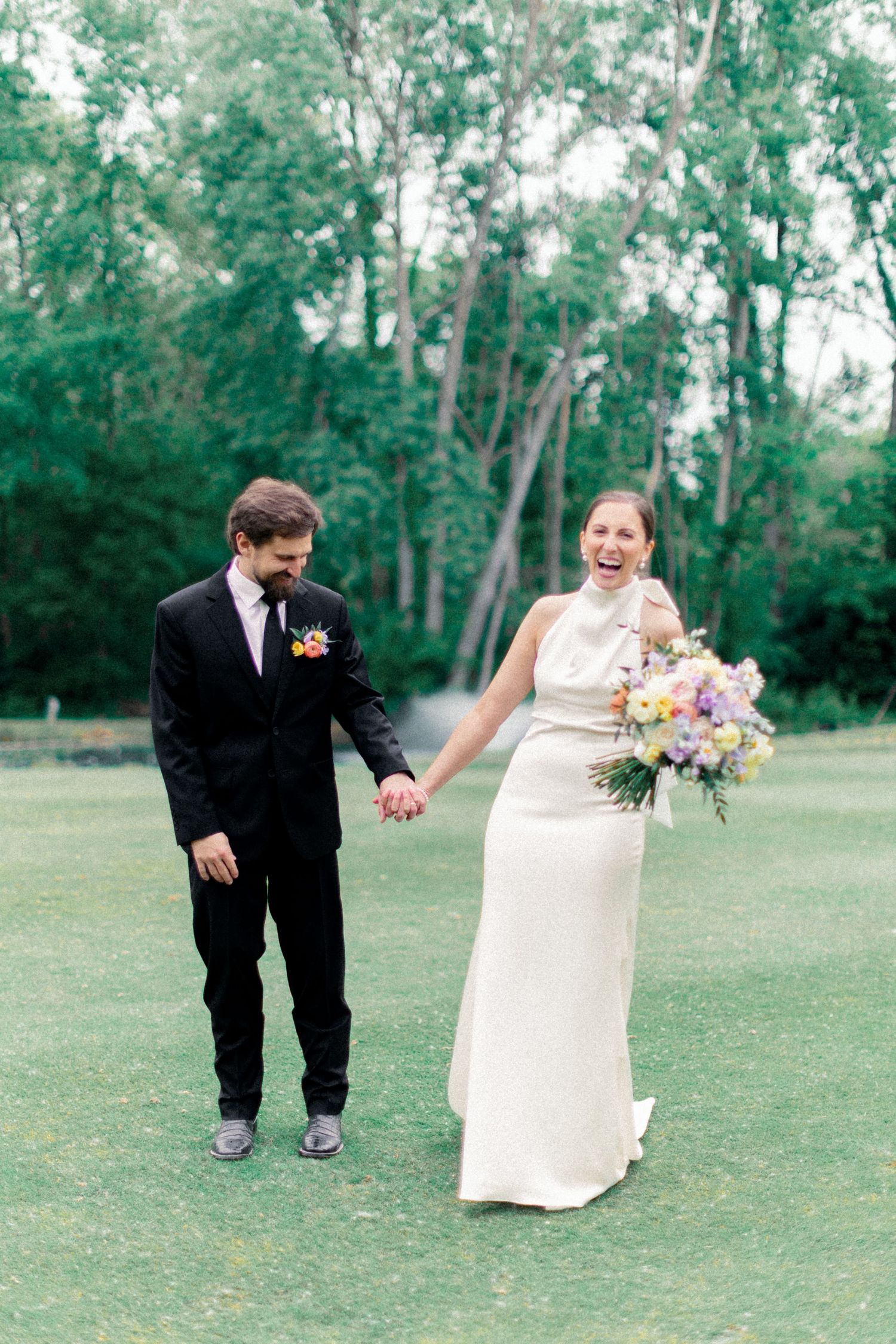 Joyful bride laughing while holding colorful bouquet as groom smiles walking hand-in-hand outdoors.
