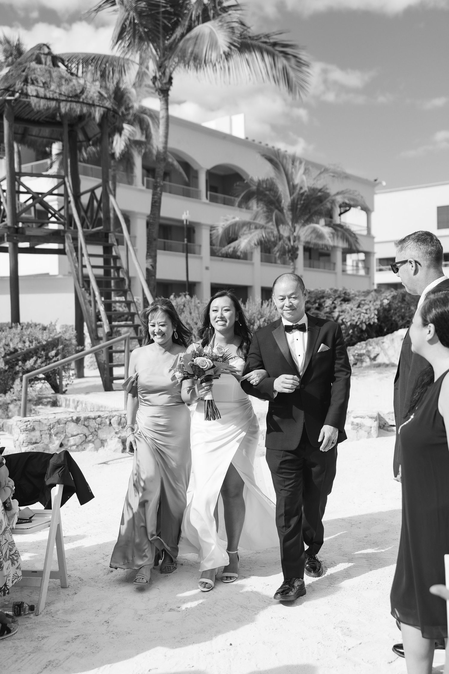 Black and white photo of a bride walking down the aisle with her parents at a beach wedding ceremony.