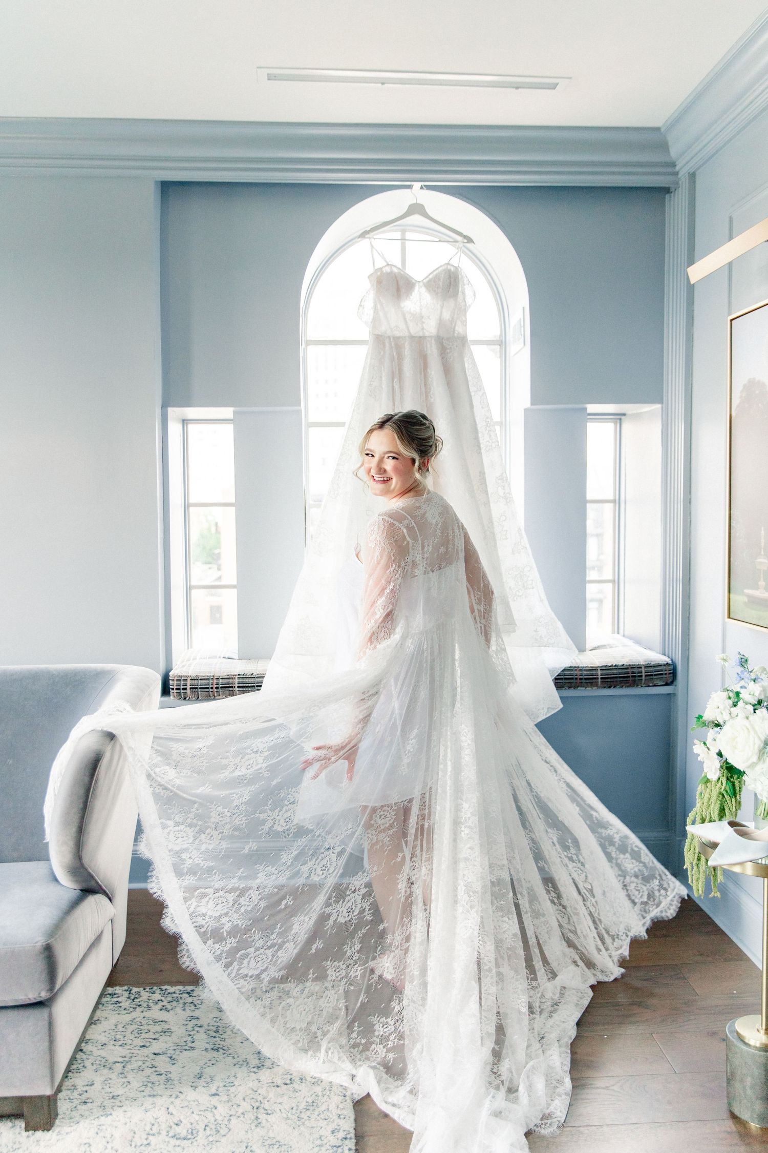 Bride in lace robe twirling near window with wedding dress hanging, in a blue bridal suite.