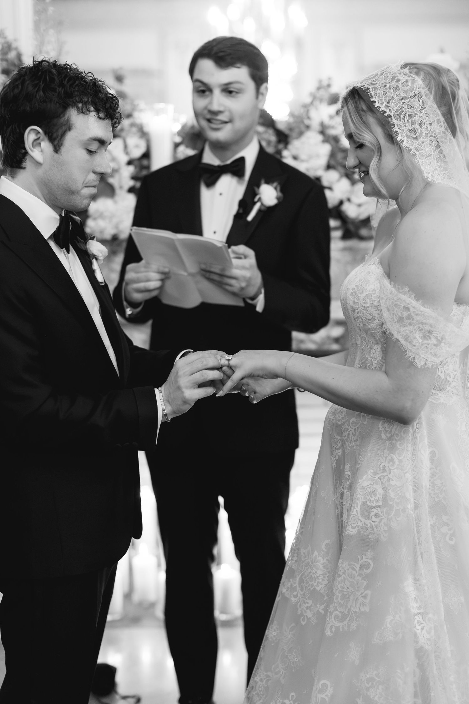 Black and white photo of groom placing wedding ring on bride's finger during ceremony with officiant.