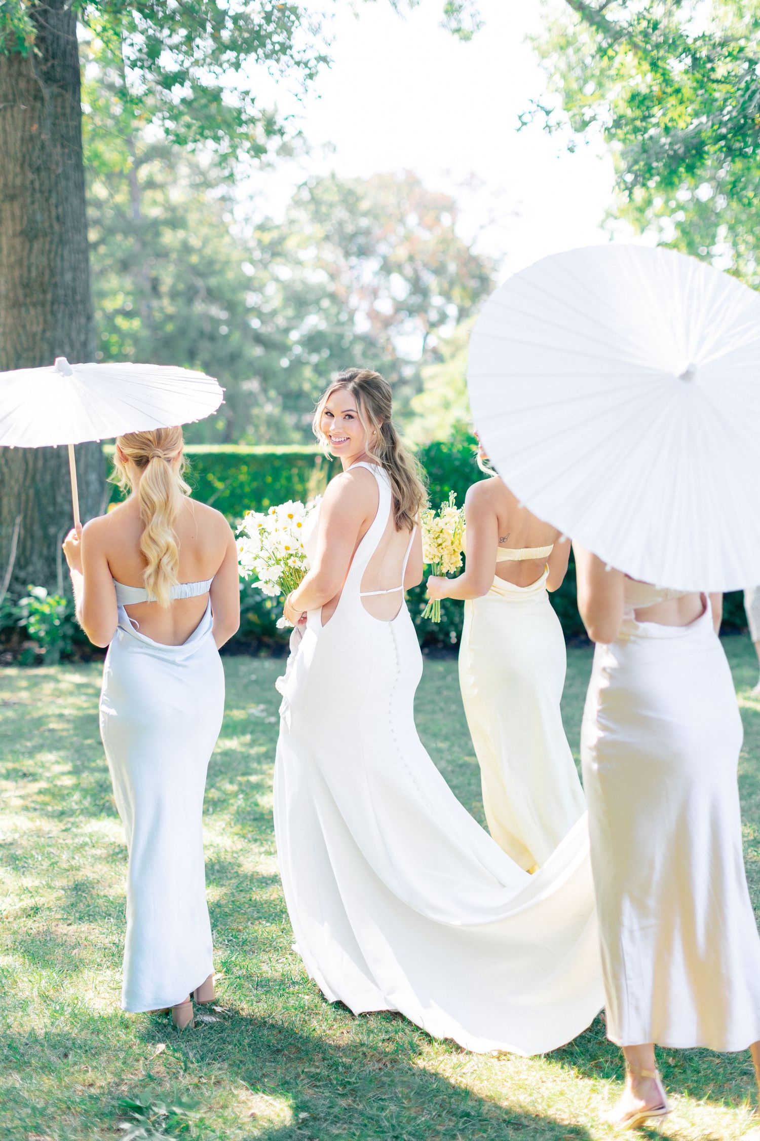 Bride in white gown smiles at camera flanked by bridesmaids in silver dresses holding white parasols outdoors.