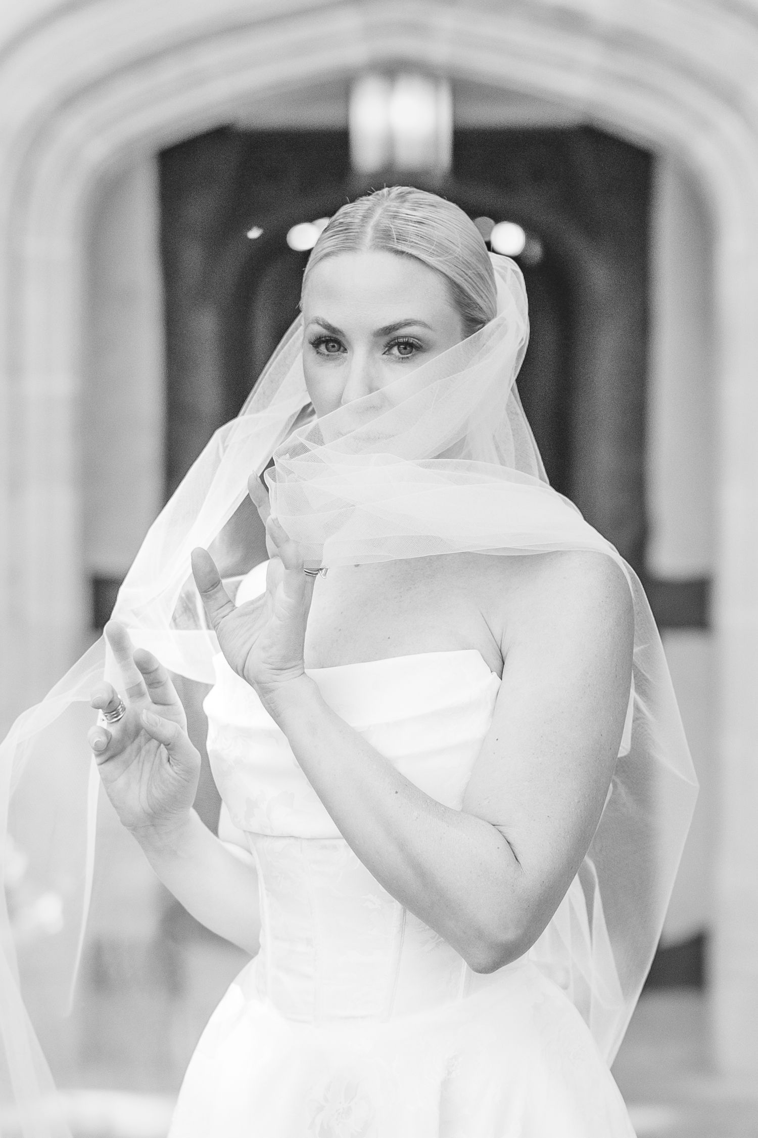 Black and white photo of a bride in a strapless gown holding her veil near her face in an elegant venue.