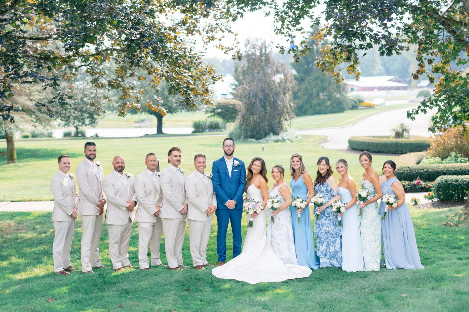 Wedding party poses outdoors, groomsmen in tan suits, bridesmaids in blue dresses, bride and groom centered on lush green lawn.