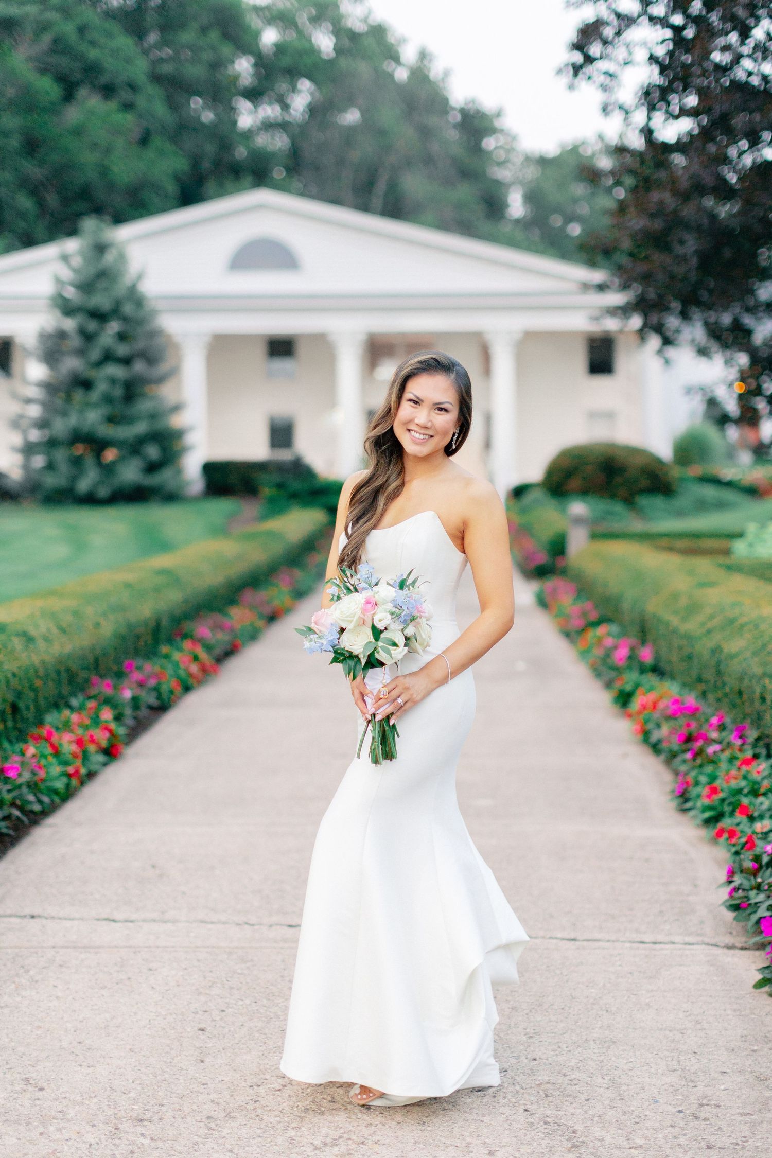 Bride in strapless white mermaid gown holding pink bouquet on garden path before elegant white mansion.