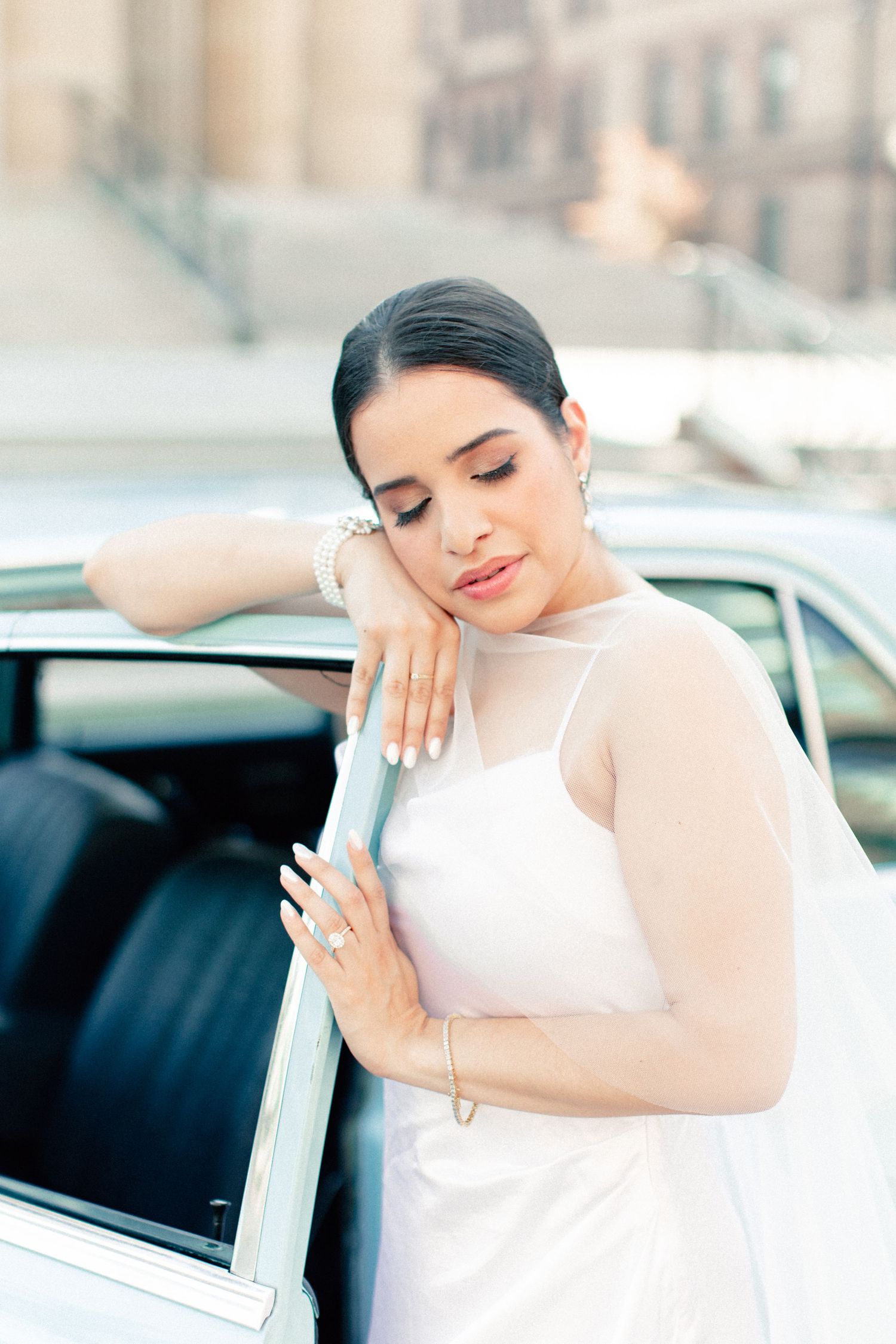 Elegant bride in white gown leaning on vintage car door, wearing pearl bracelet and veil, eyes closed.