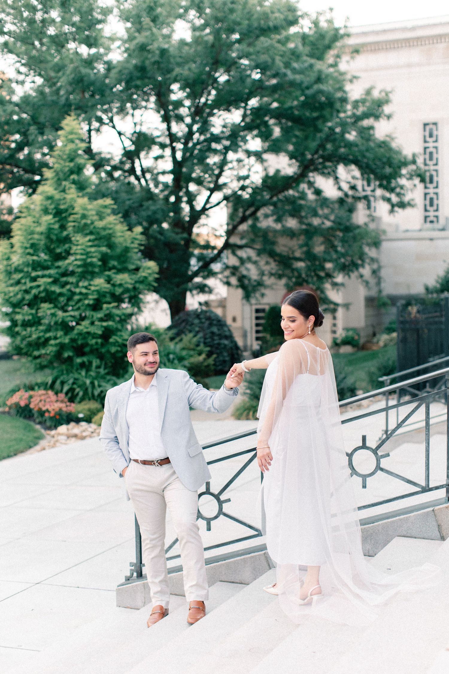 Bride in a flowing white gown leads groom by the hand up stone steps near a classic historic building.