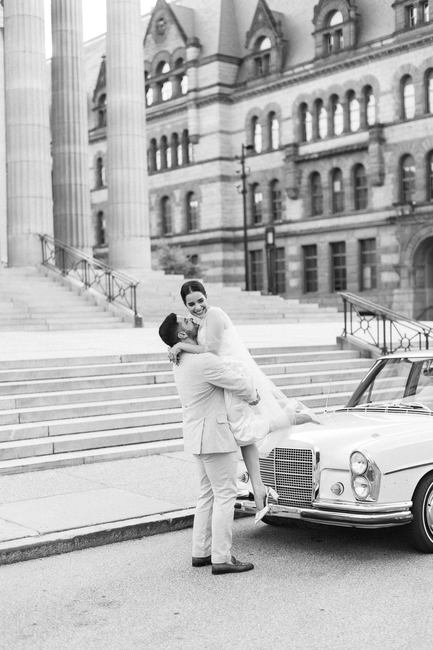 Groom lifting bride in front of historic building beside vintage Mercedes in black and white wedding photo.