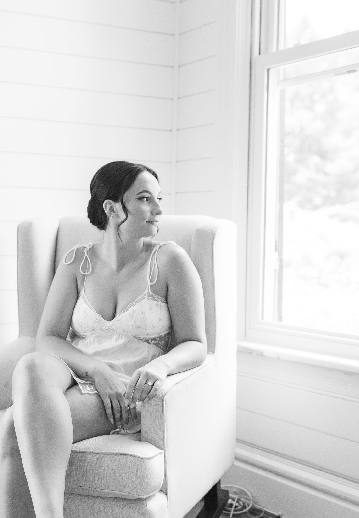 Black and white photo of a woman in lingerie sitting in a white armchair gazing out a bright window.