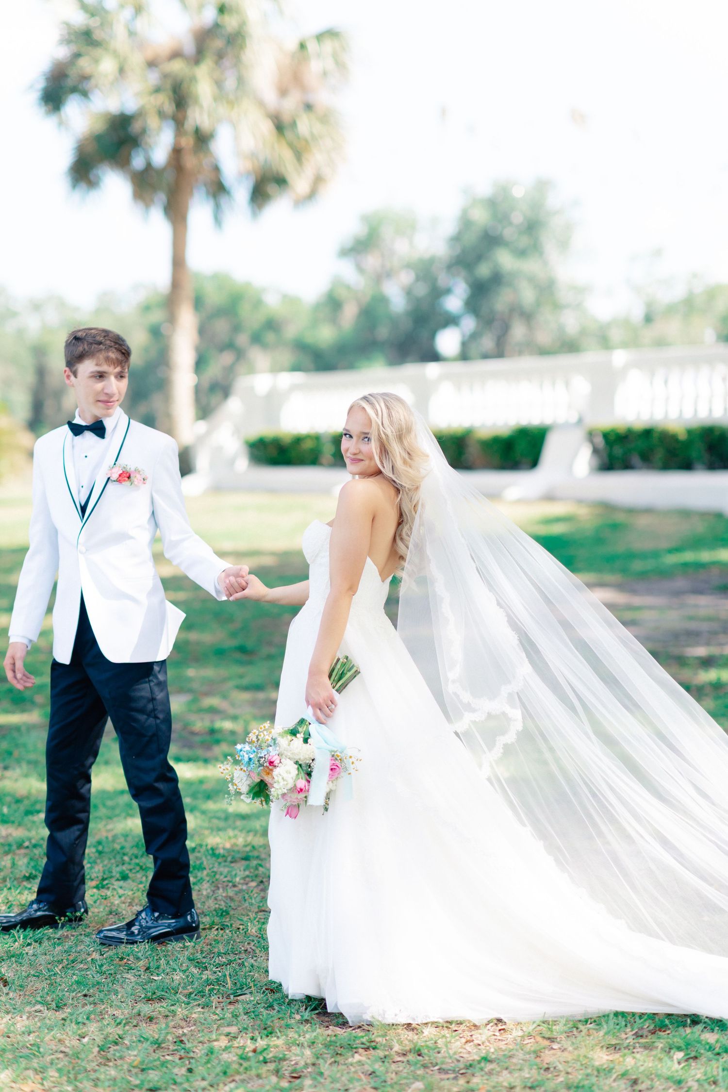 Bride in white gown with flowing veil holds bouquet while groom in white tuxedo holds her hand outdoors, wedding at Bella Cosa