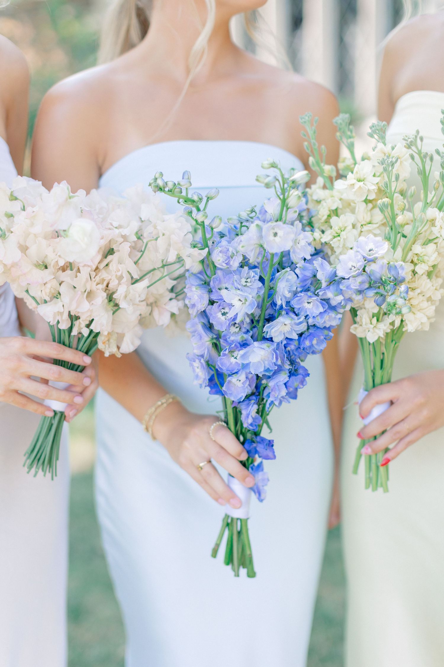 Three bridesmaids in light blue dresses holding bouquets of blue delphinium and white stock flowers outdoors.