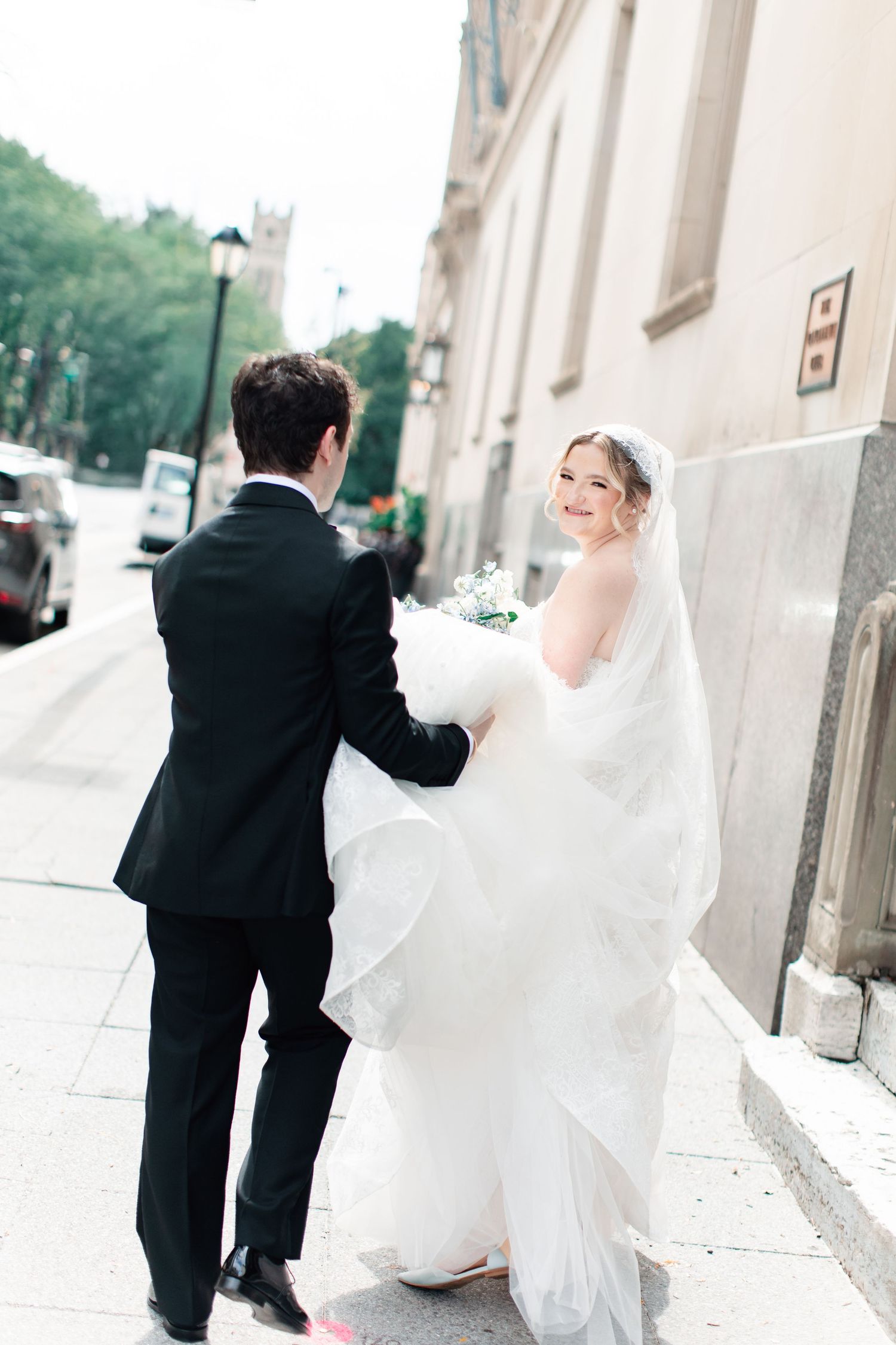 Bride in white gown and veil walks with groom in black tuxedo along urban sidewalk, smiling over her shoulder.
