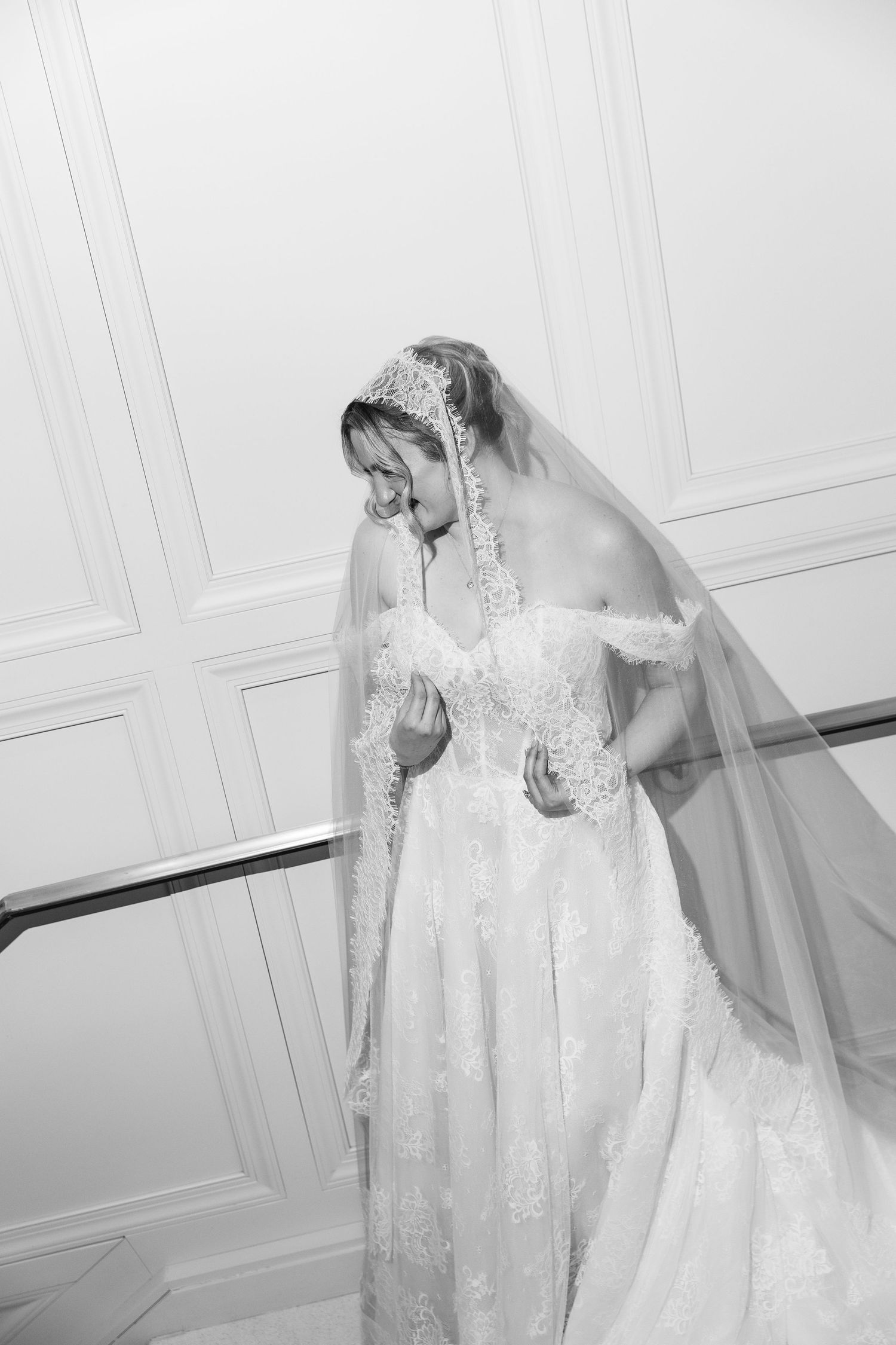 Black and white photo of a bride in lace off-shoulder gown and veil, looking down gracefully on staircase.