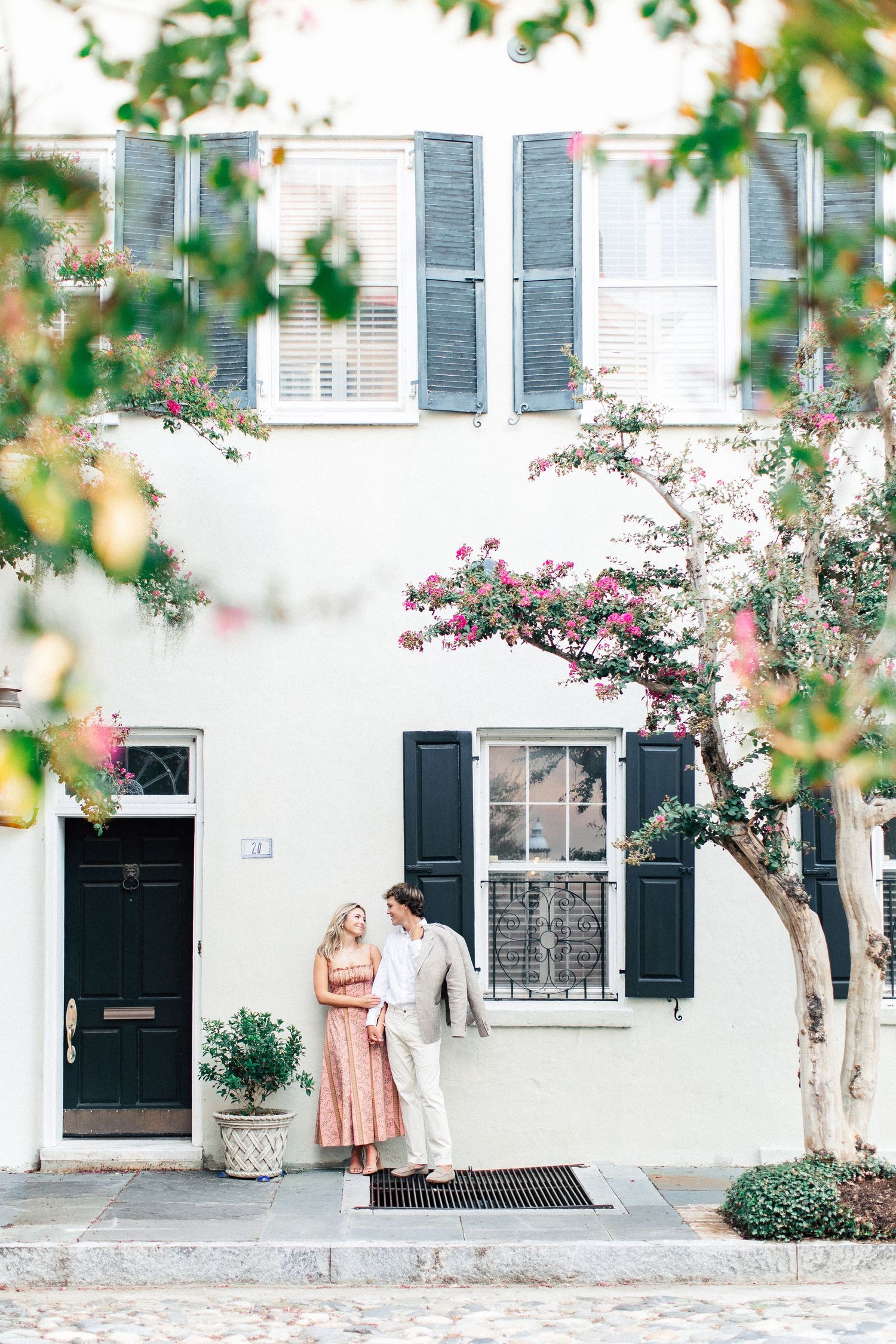 Couple standing together outside a white historic Charleston home with black shutters and blooming pink crape myrtle trees.