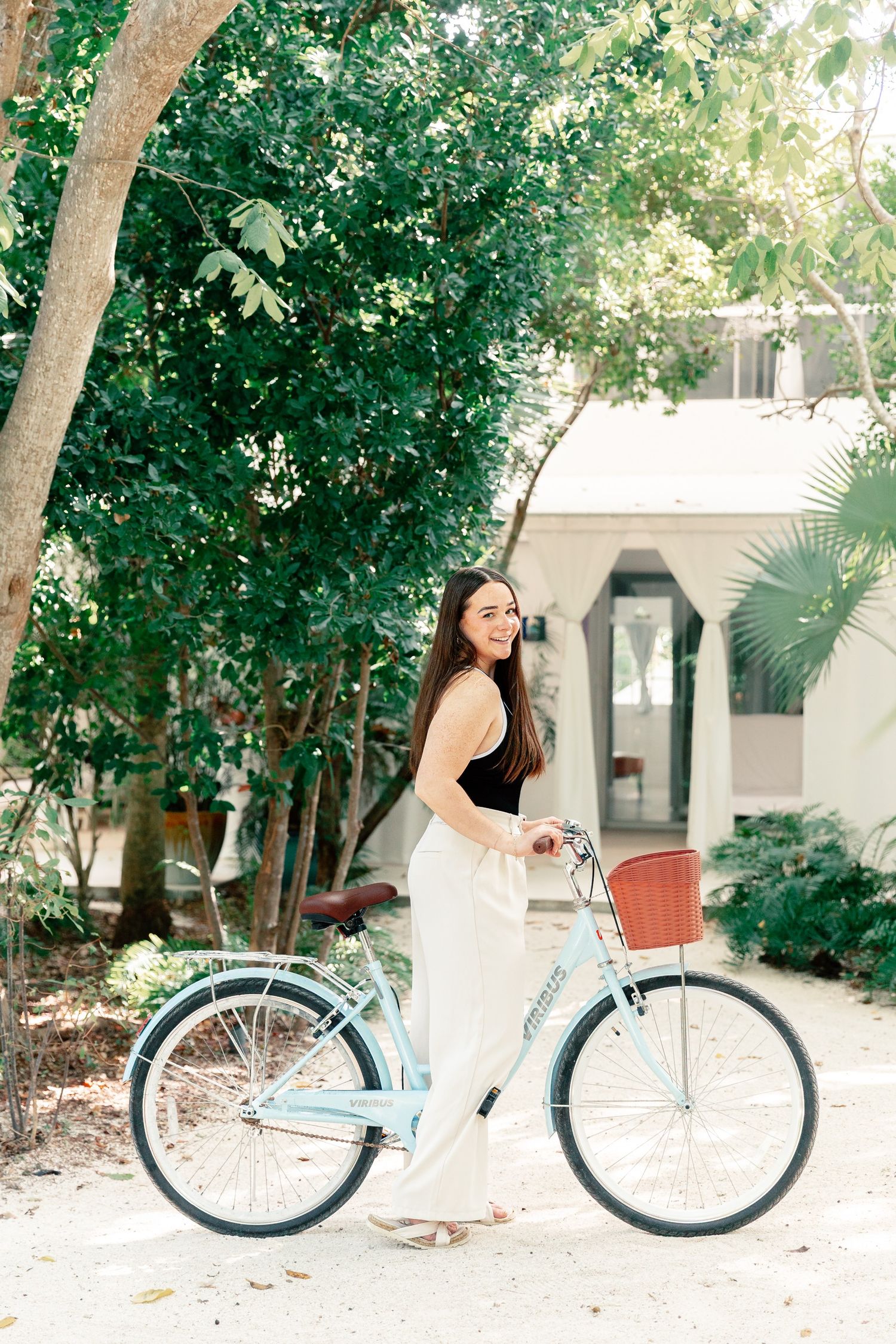 Young woman smiling beside a light blue cruiser bicycle with a basket, surrounded by lush green trees.