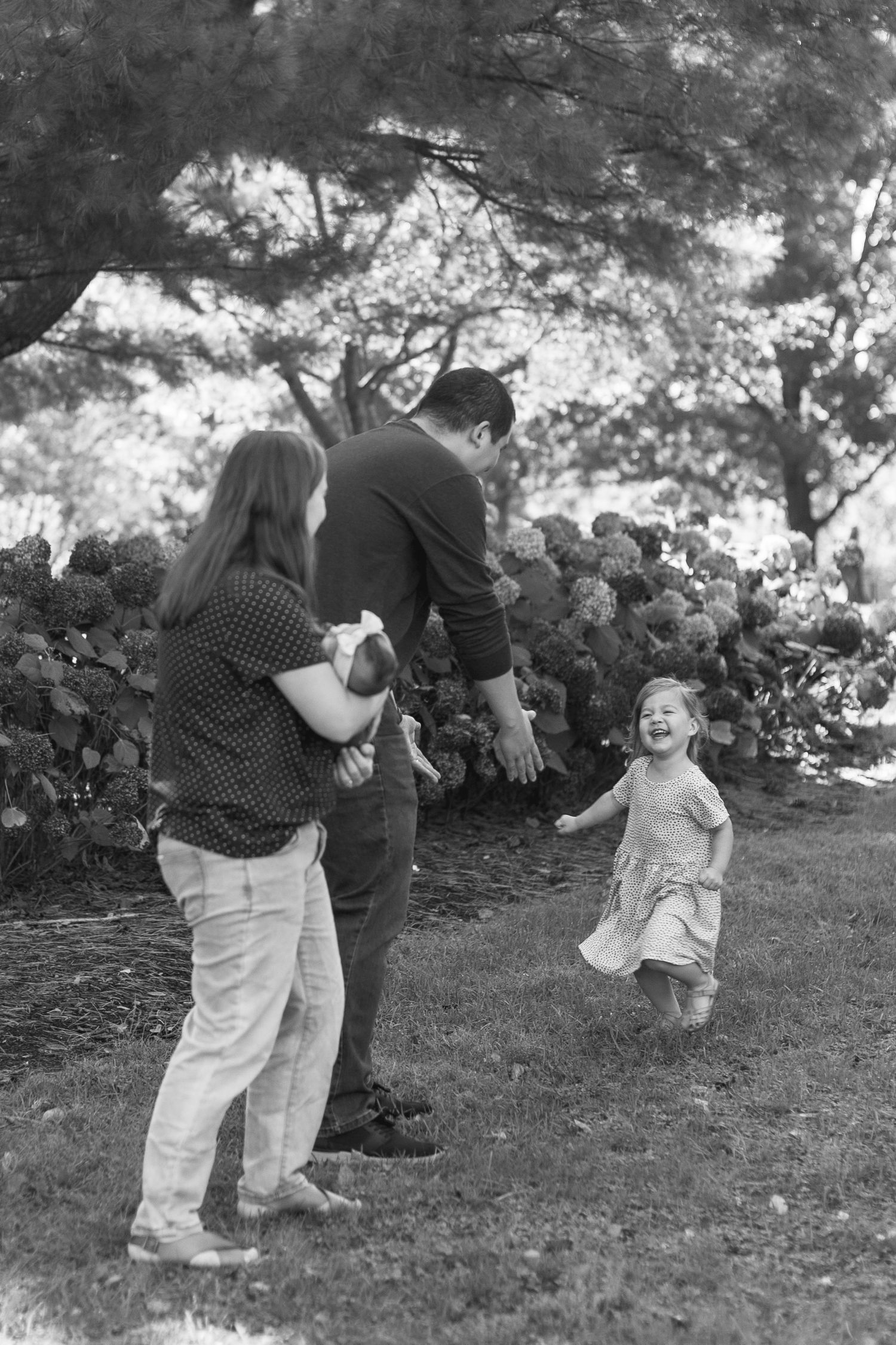 Black and white photo of a joyful toddler running toward her parents in a lush garden setting.