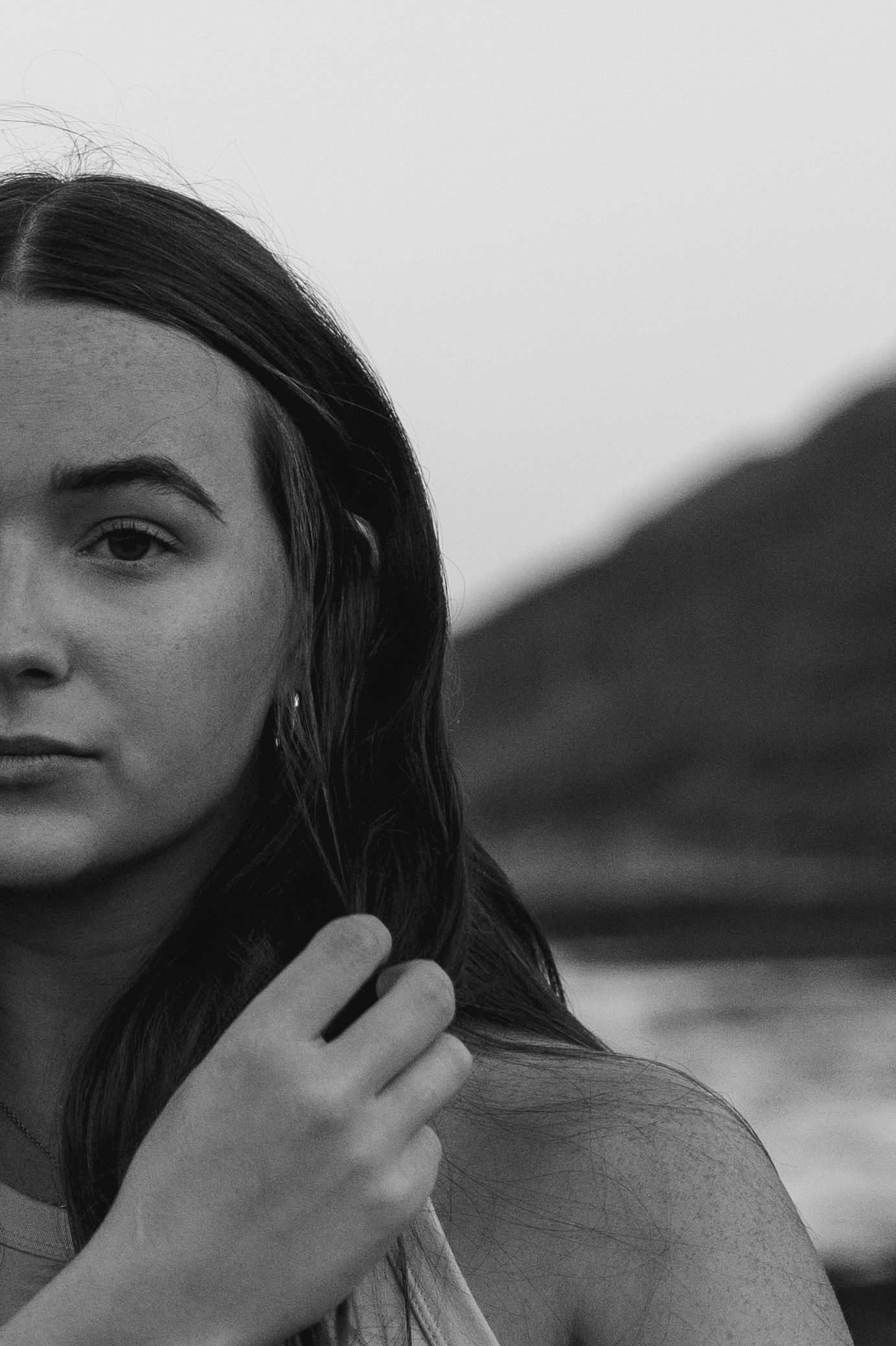 Black and white portrait of a young woman with long hair holding her hair back, mountains in background.