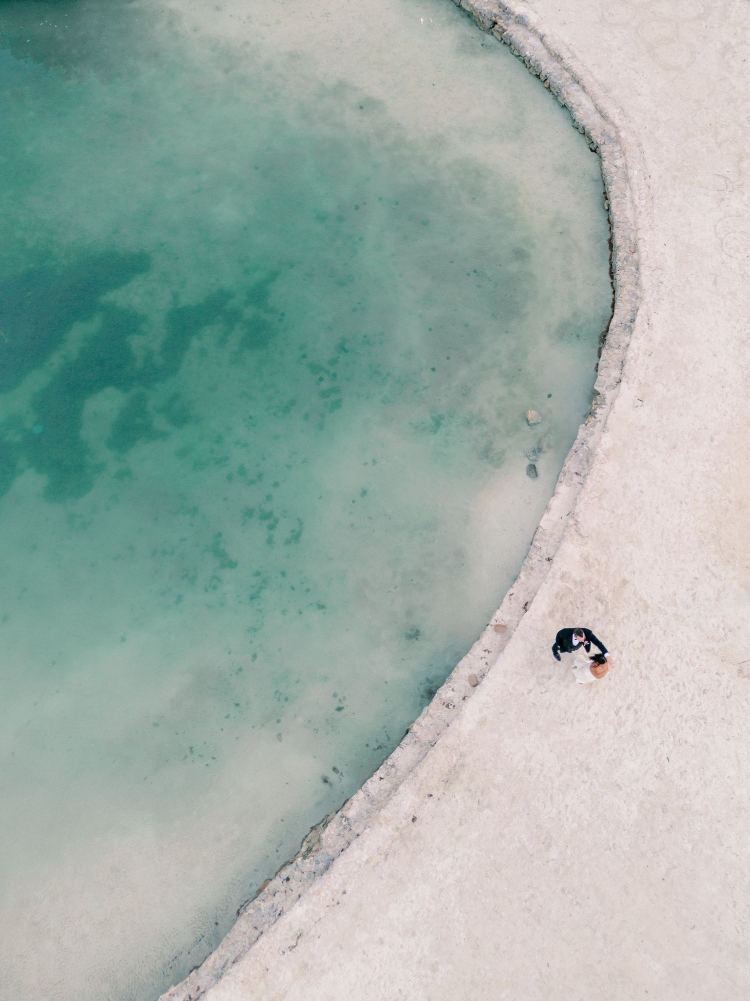 Aerial drone view of couple in wedding attire sitting beside a stunning turquoise natural pool.