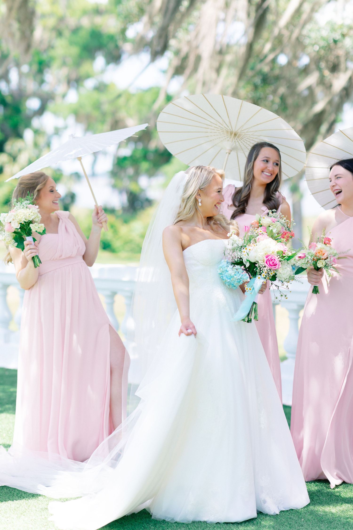 Bride in white gown with bridesmaids in pink dresses holding parasols and floral bouquets at outdoor wedding.