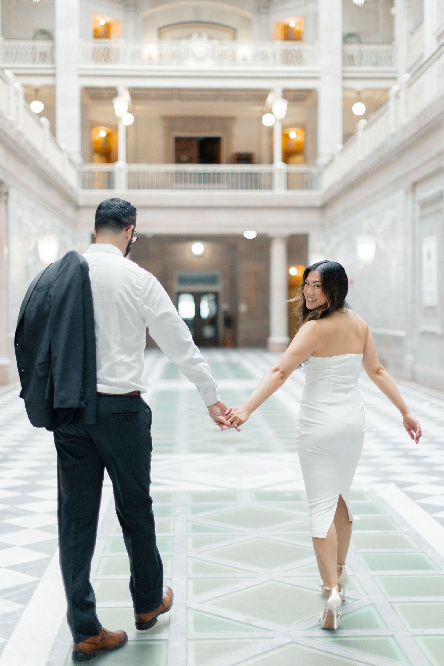 Couple holding hands walking through elegant marble hall, woman in white strapless dress, man in dark suit jacket over shoulder.