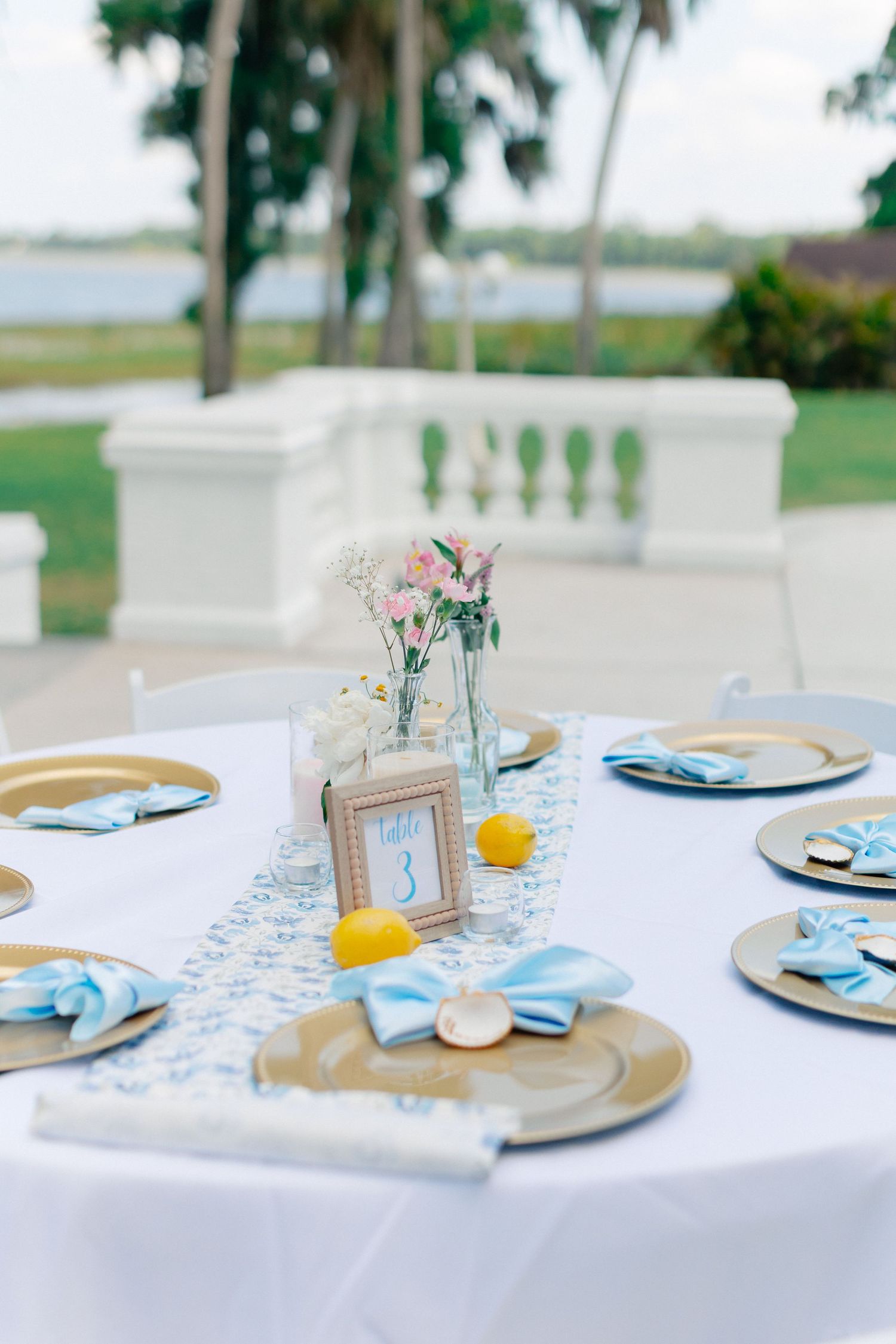Elegant outdoor wedding reception table with gold chargers, blue napkins, lemon centerpieces, and floral arrangements on white tablecloth