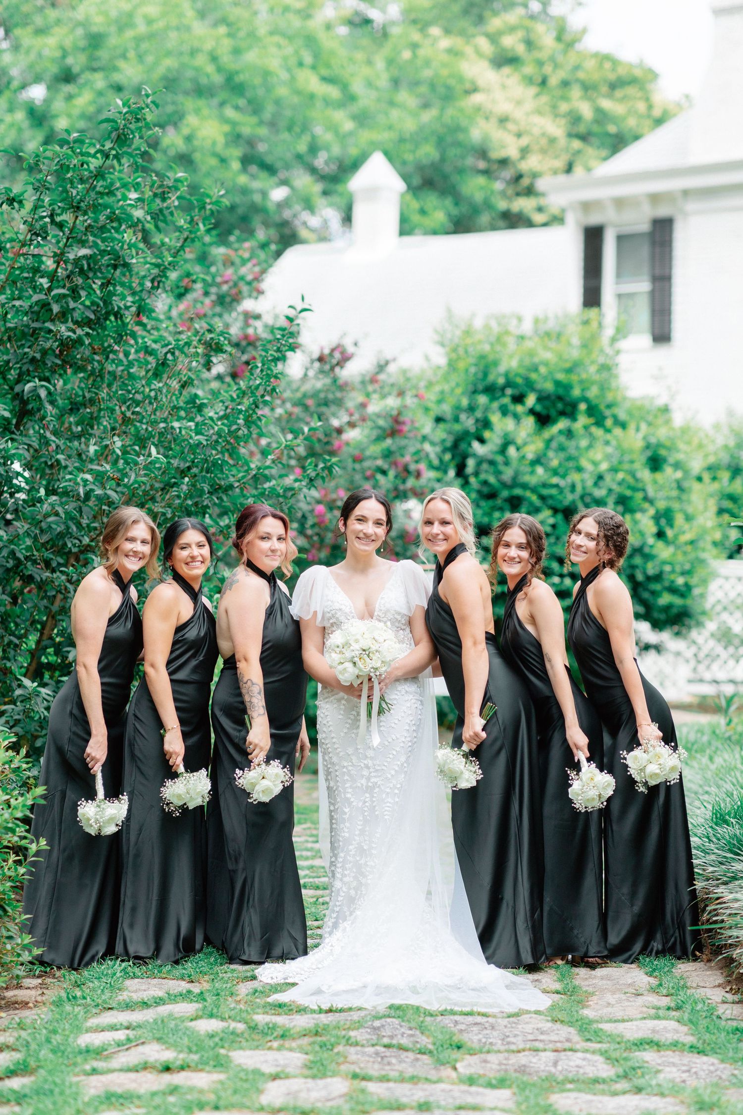 Bride in white lace gown poses with six bridesmaids in black dresses holding white bouquets in a garden.