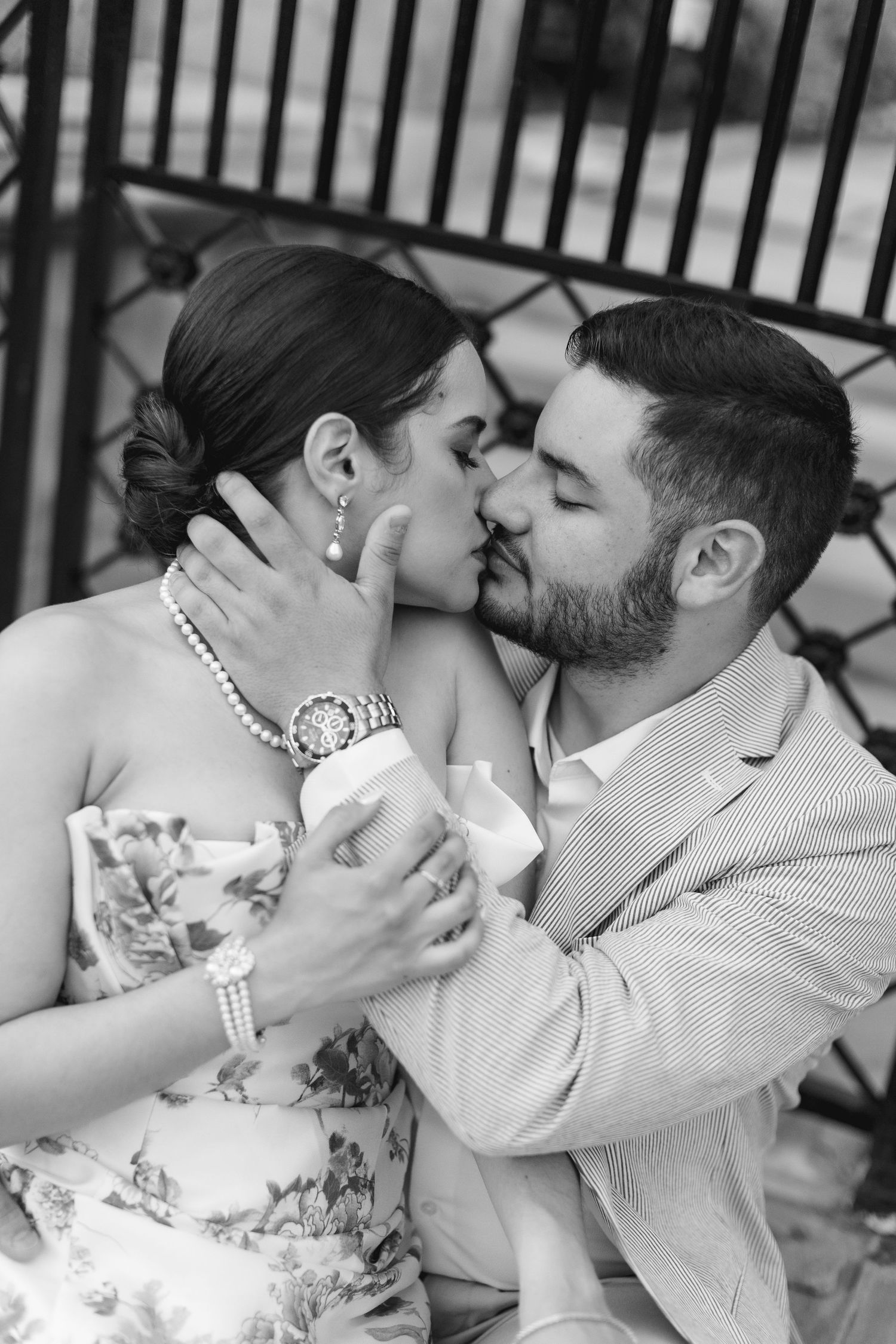 Black and white photo of a couple sharing a romantic kiss, woman wearing pearl necklace and floral dress.