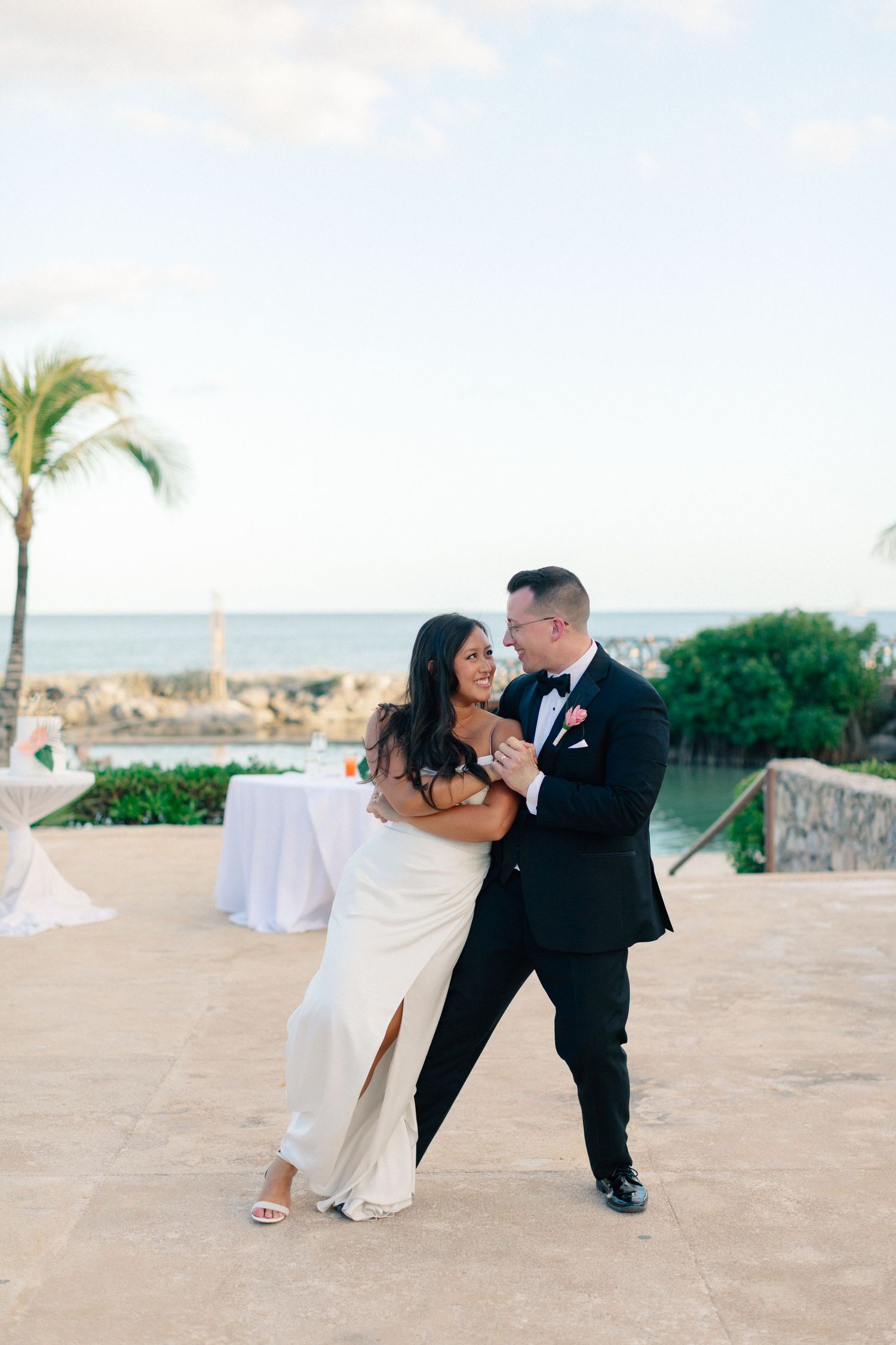 Bride and groom share their first dance at an outdoor oceanfront wedding reception, palm trees and sea in the background.
