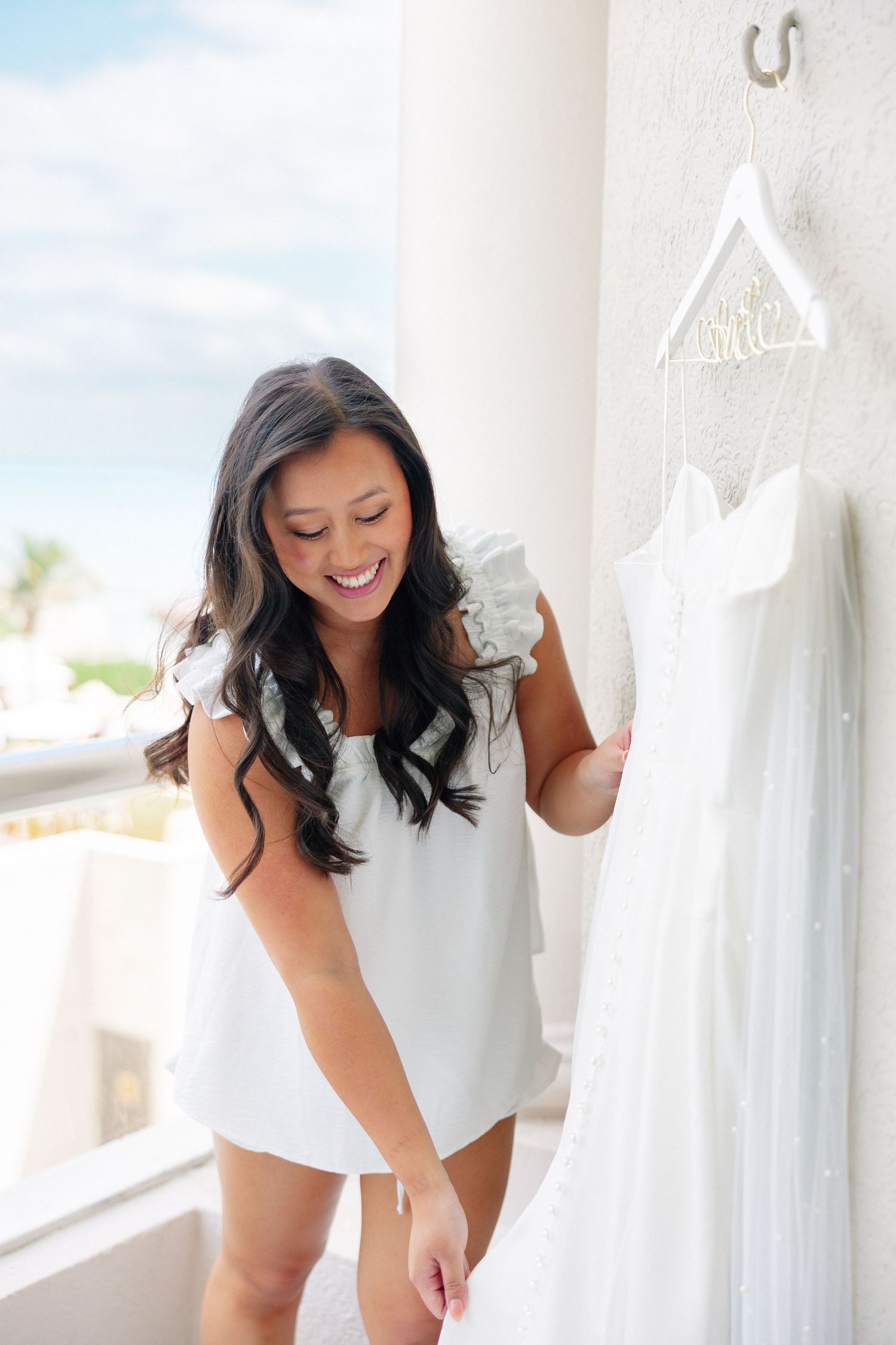 Smiling bride in white top admiring her wedding dress hanging on a decorative hanger near a bright window.