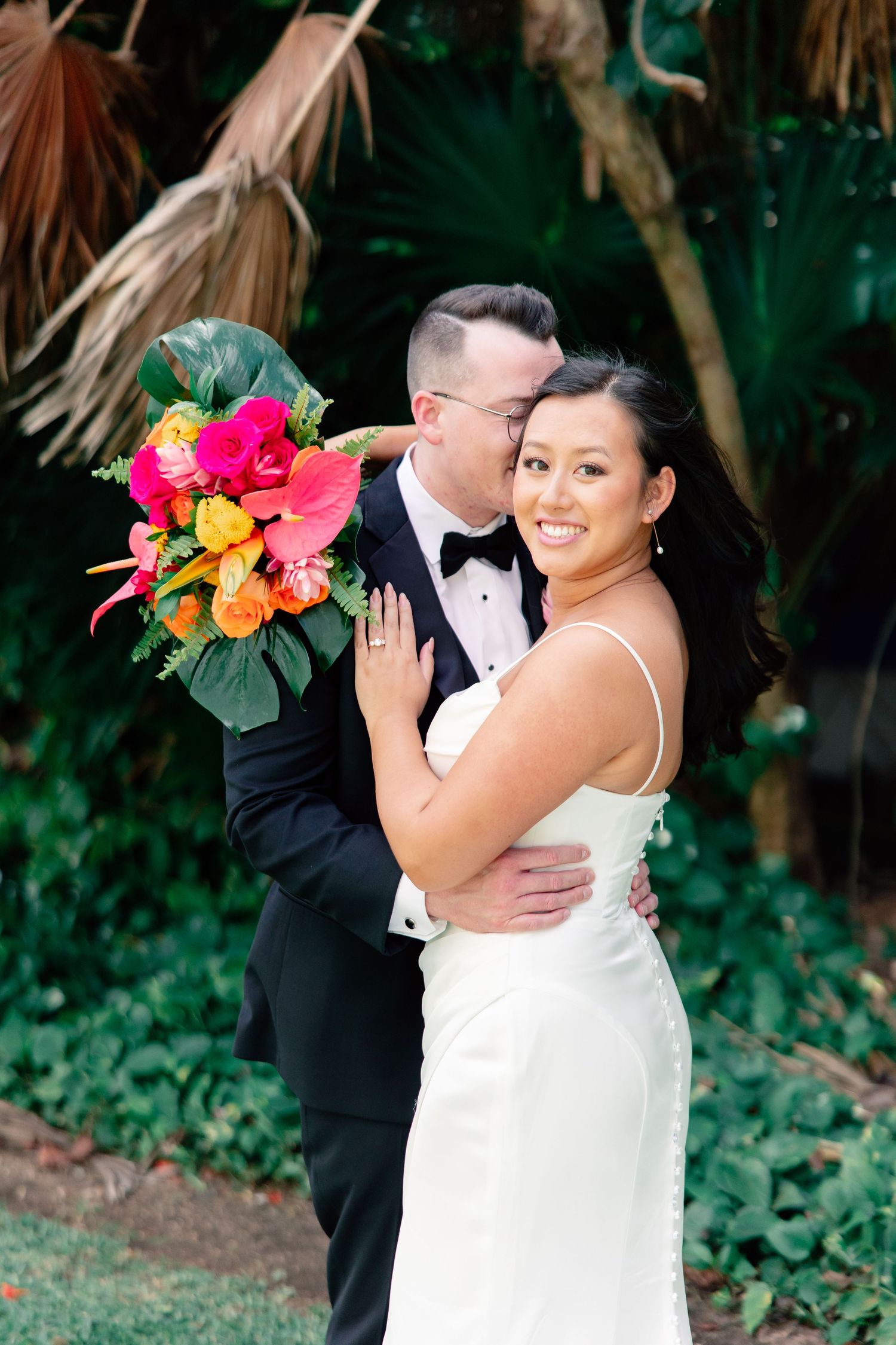 Bride in white gown holds vibrant tropical bouquet while groom in tuxedo embraces her outdoors.