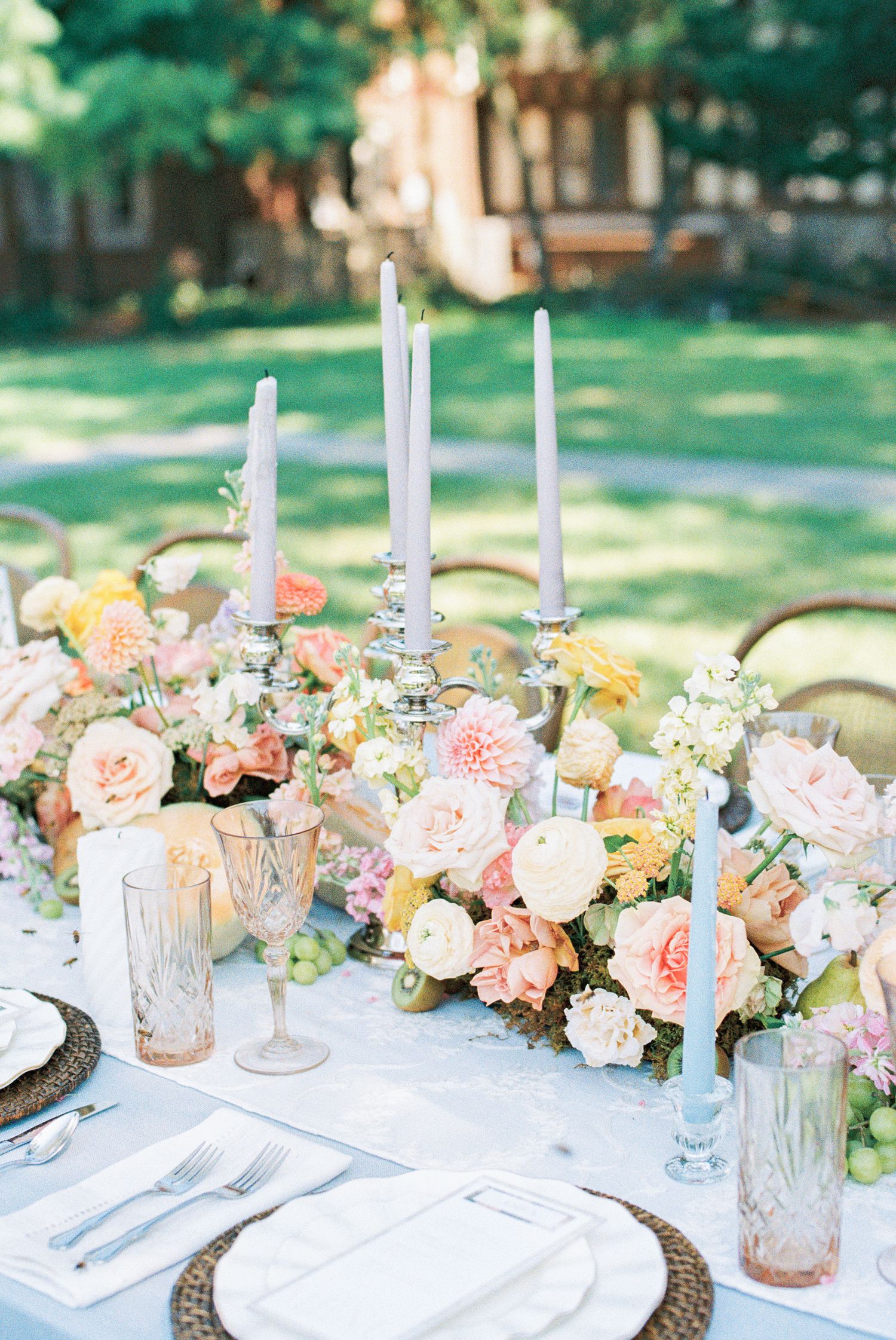Elegant outdoor wedding tablescape with pastel floral centerpiece, taper candles, woven chargers, and colorful glassware on white linen.