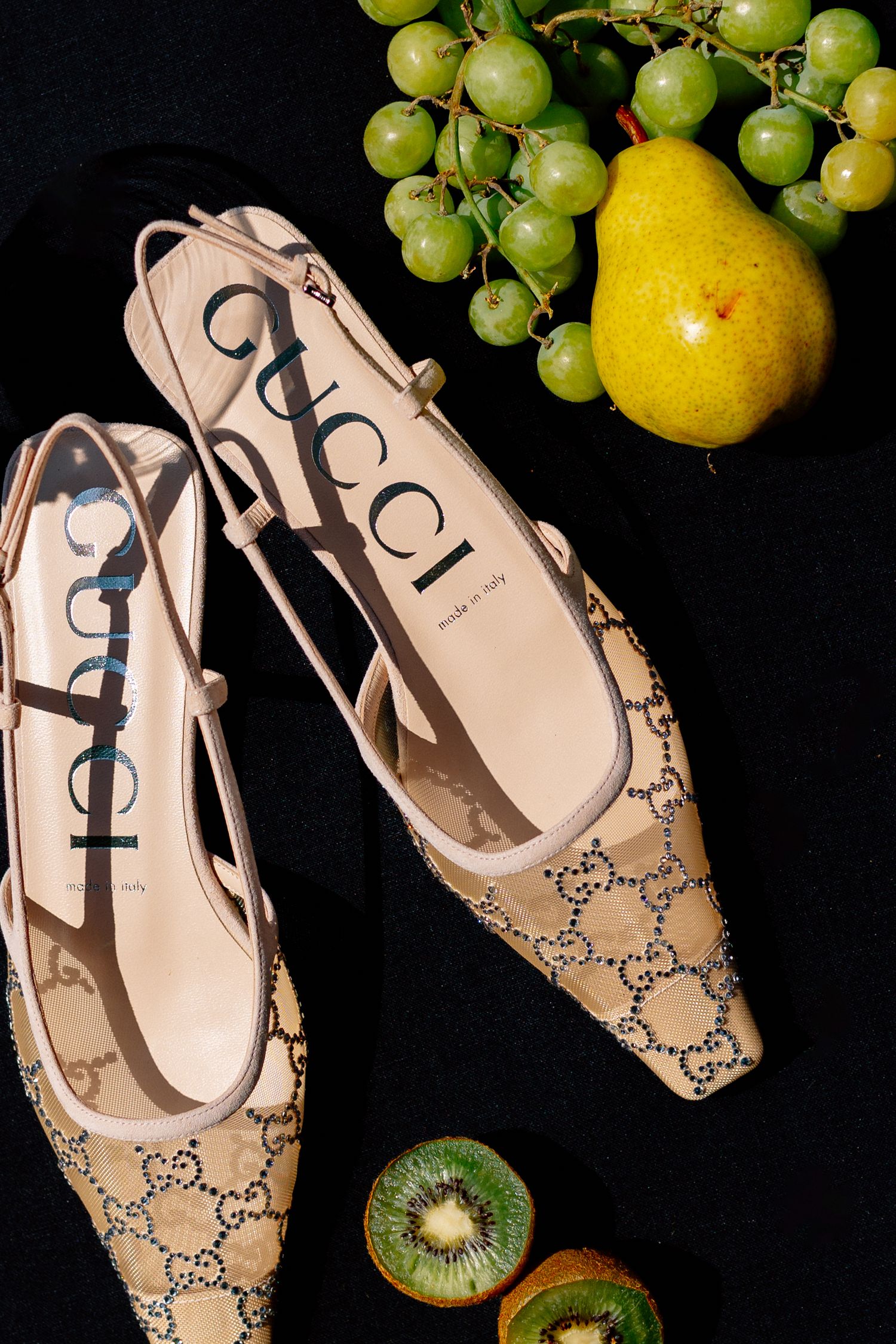 Gucci beige monogram slingback heels displayed on black surface with fresh green grapes, pear, and kiwi fruit.