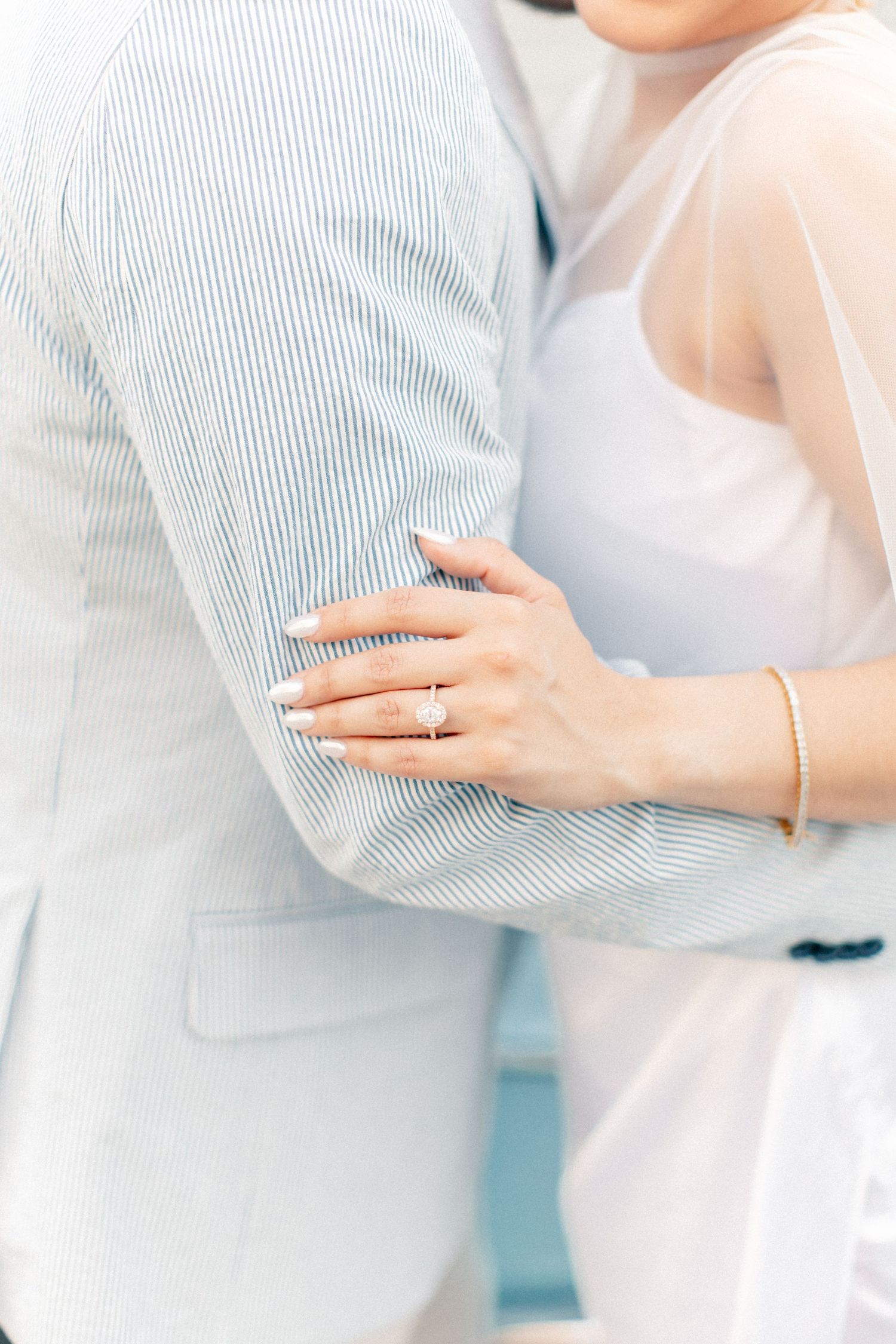 Close-up of bride's hand with engagement ring and bracelet embracing groom in striped blue seersucker suit.