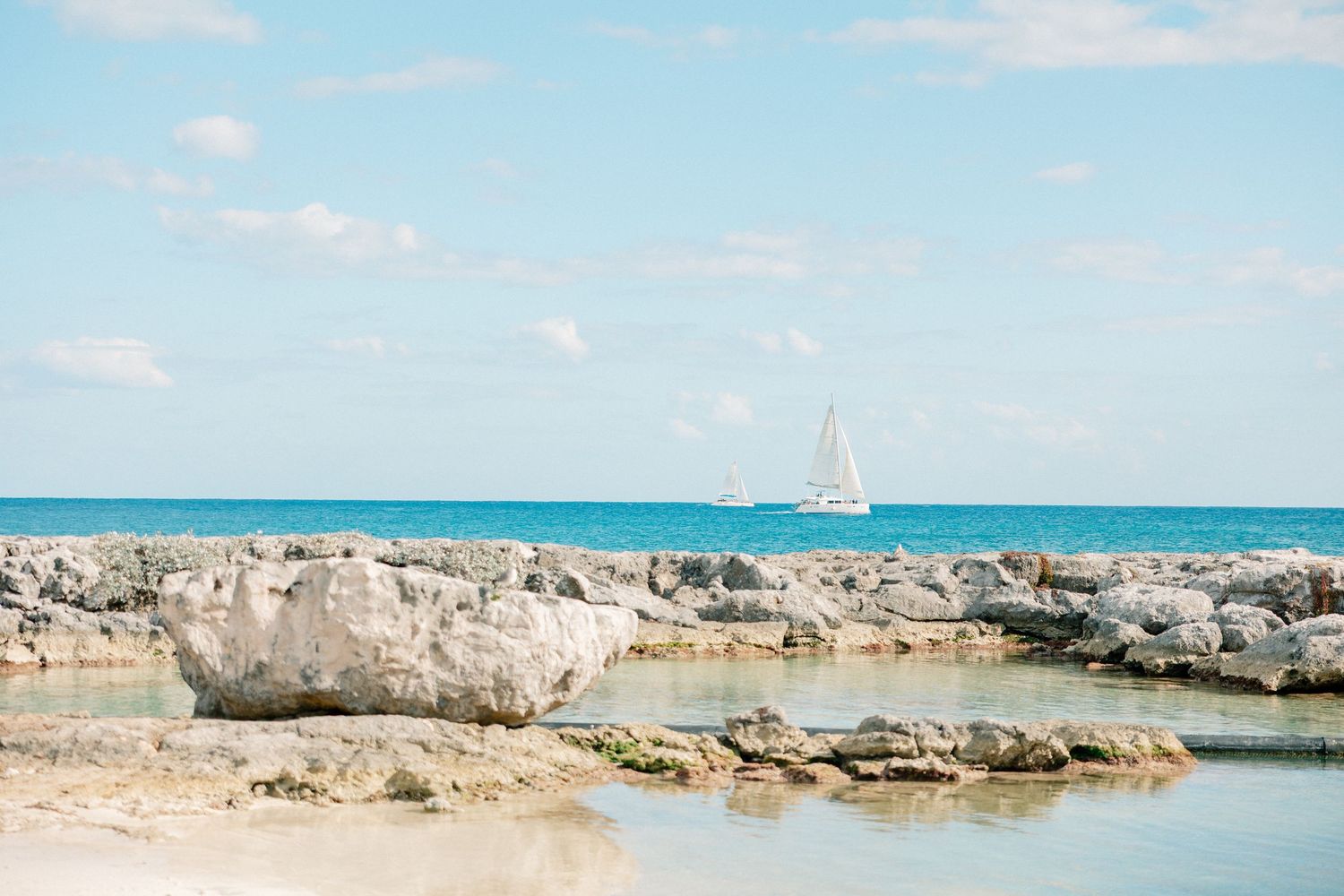 Rocky shoreline with crystal-clear turquoise water and a white sailboat sailing on the calm blue sea under a partly cloudy sky.