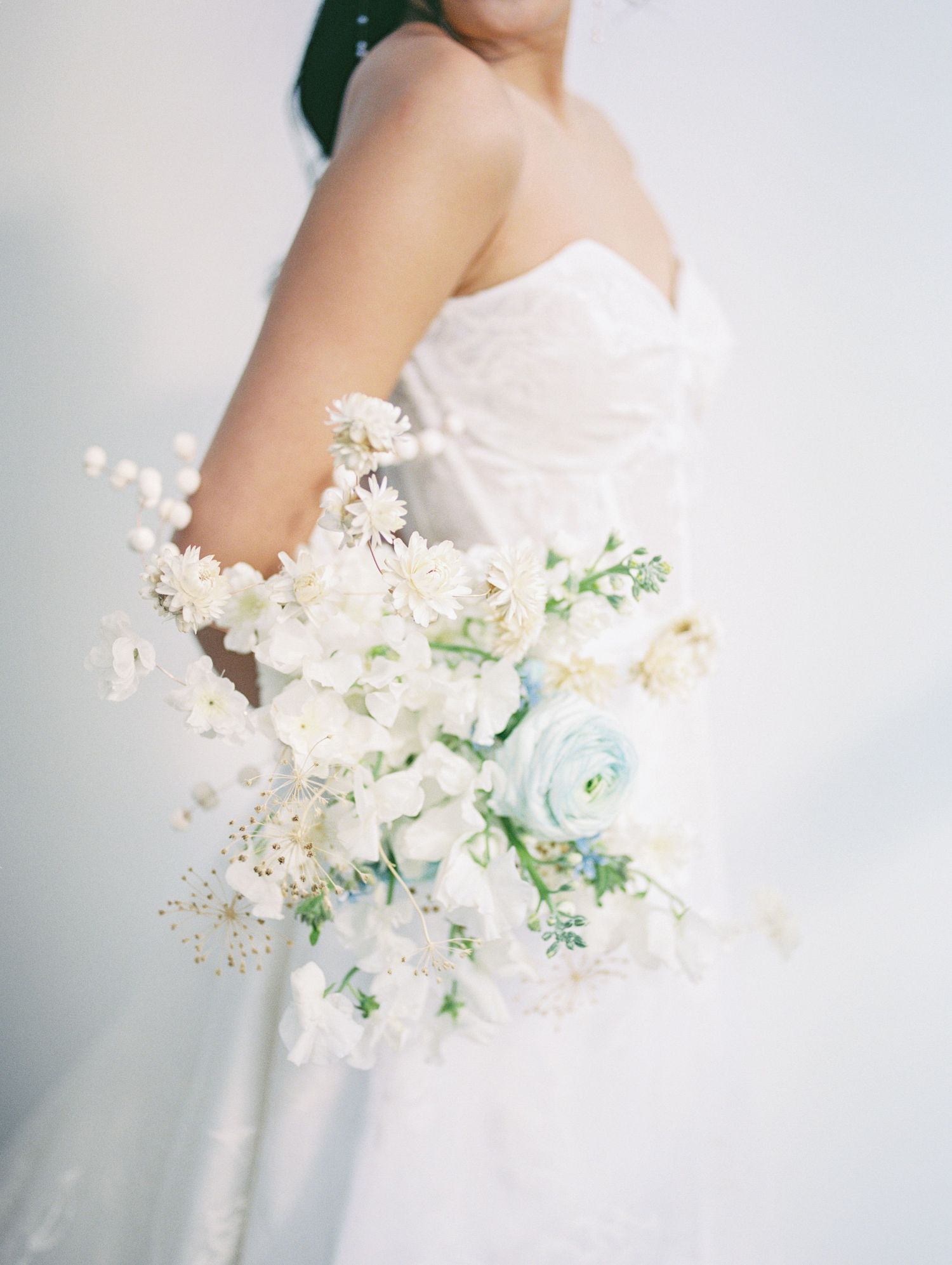 Bride in white strapless gown holding a soft bouquet of white and pale blue flowers including ranunculus and daisies.