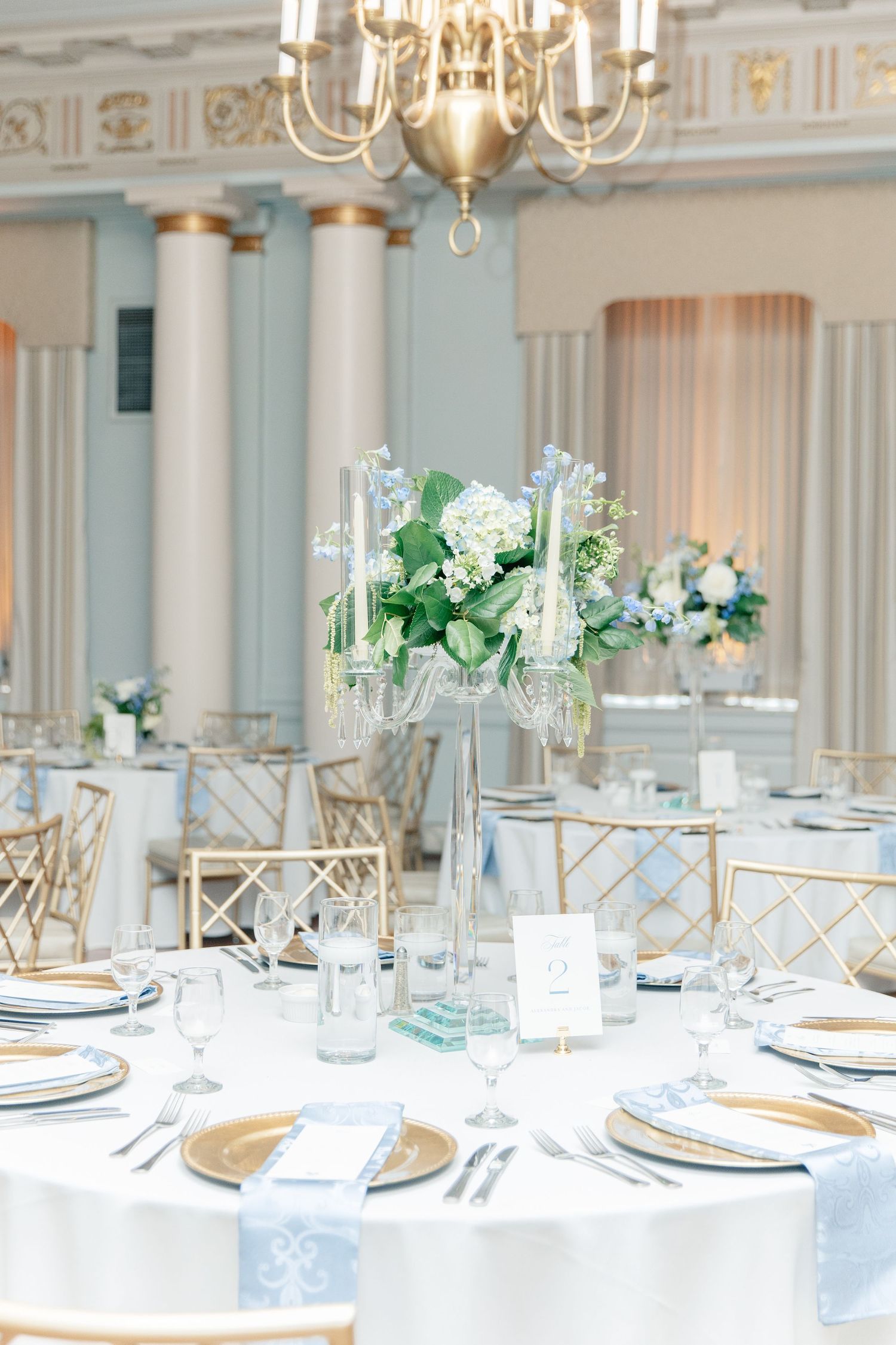 Elegant wedding reception table with tall floral centerpiece, gold chargers, crystal glasses, and blue accents in a grand ballroom.