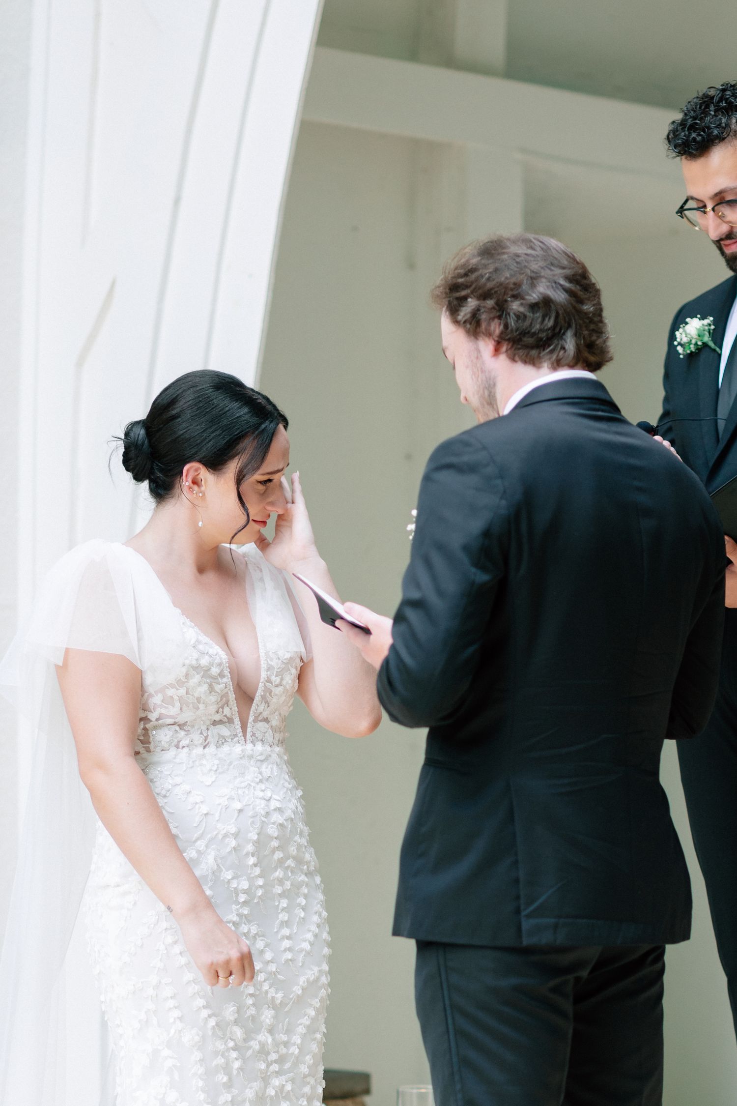 Bride wiping tears as groom reads wedding vows during emotional outdoor ceremony, wearing beaded white gown.