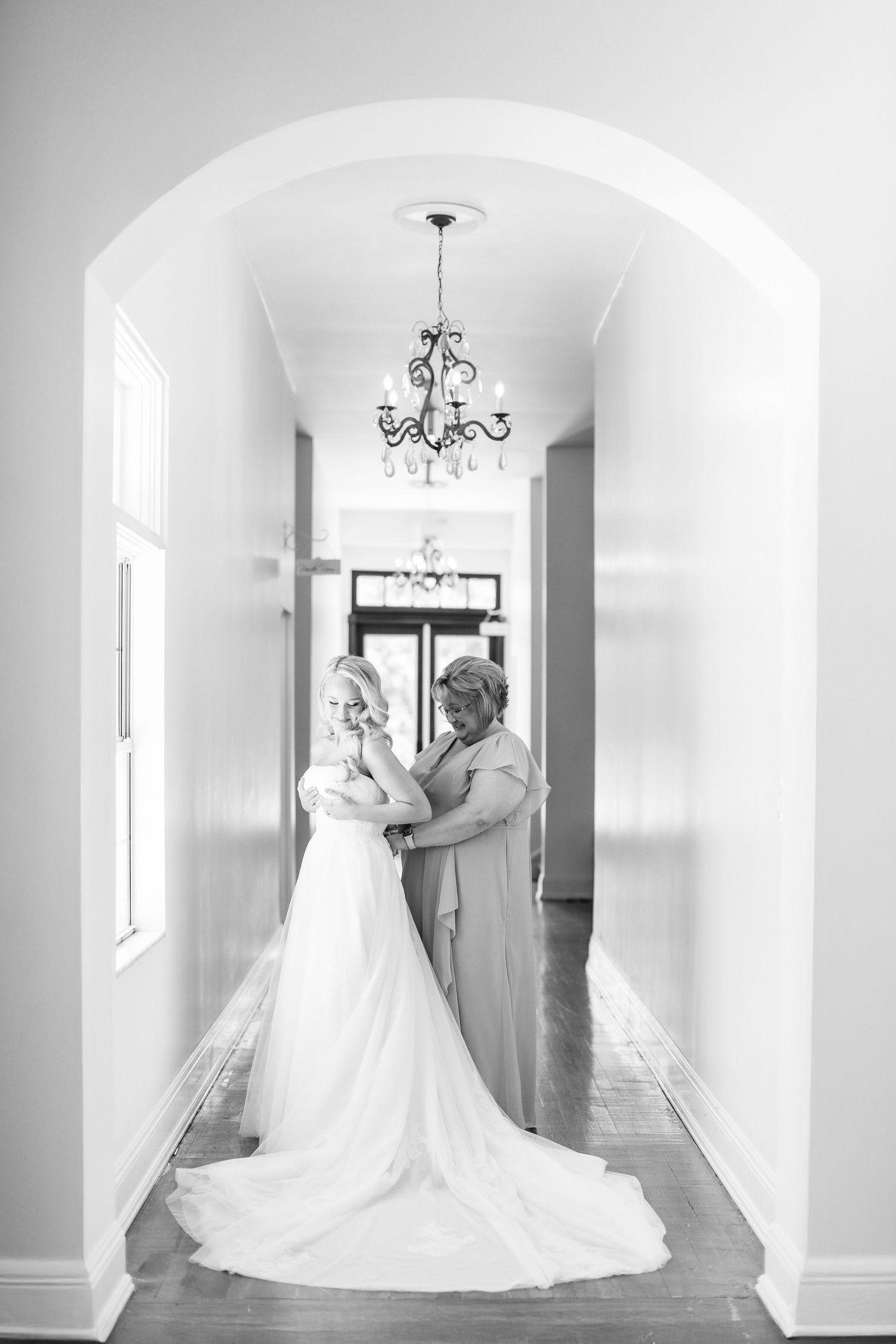 Black and white photo of a bride in a wedding gown being helped by her mother in an elegant hallway, wedding at Bella Cosa