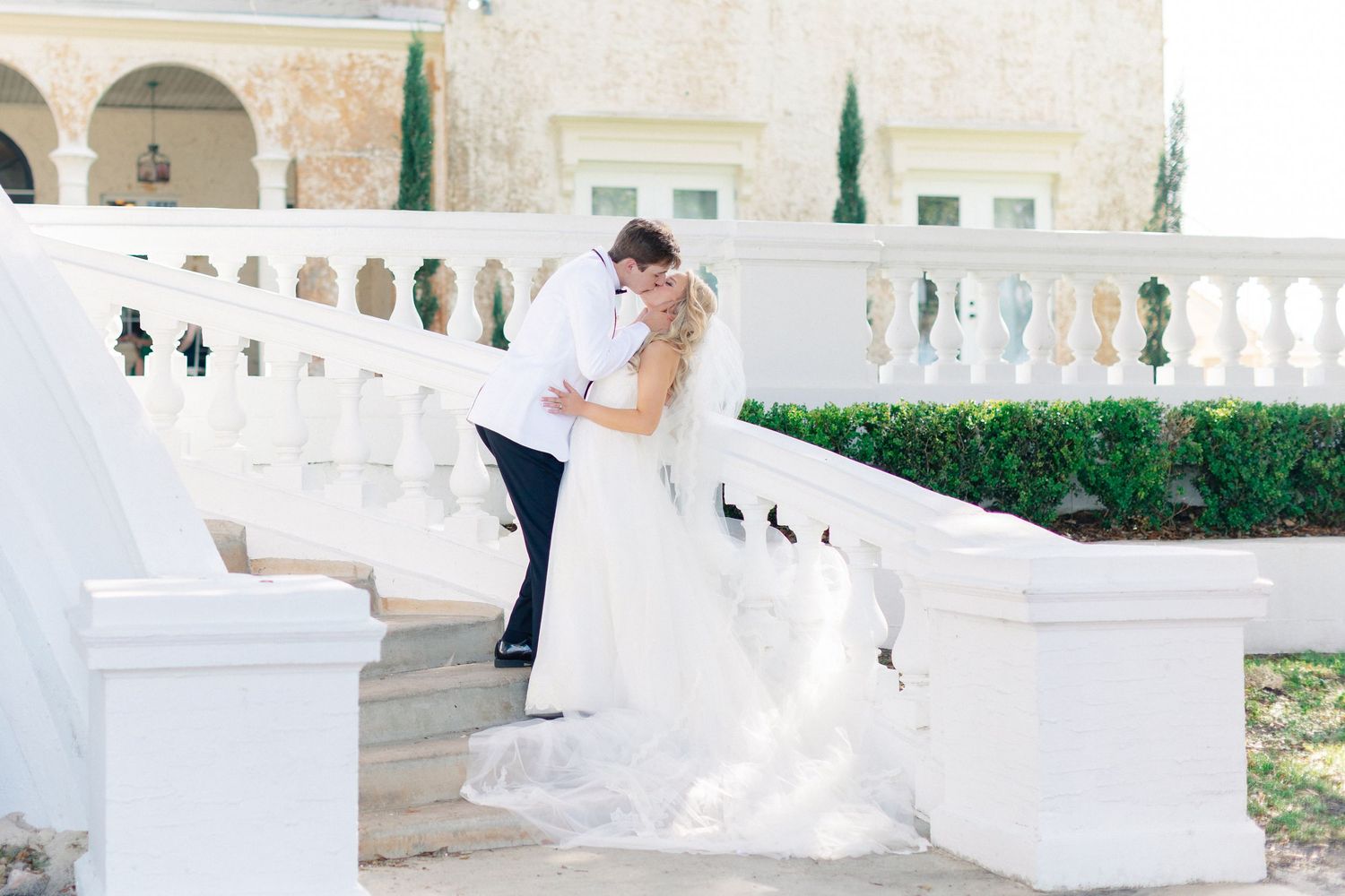 Bride and groom share a romantic kiss on white stone steps outside an elegant mansion venue, wedding at Bella Cosa