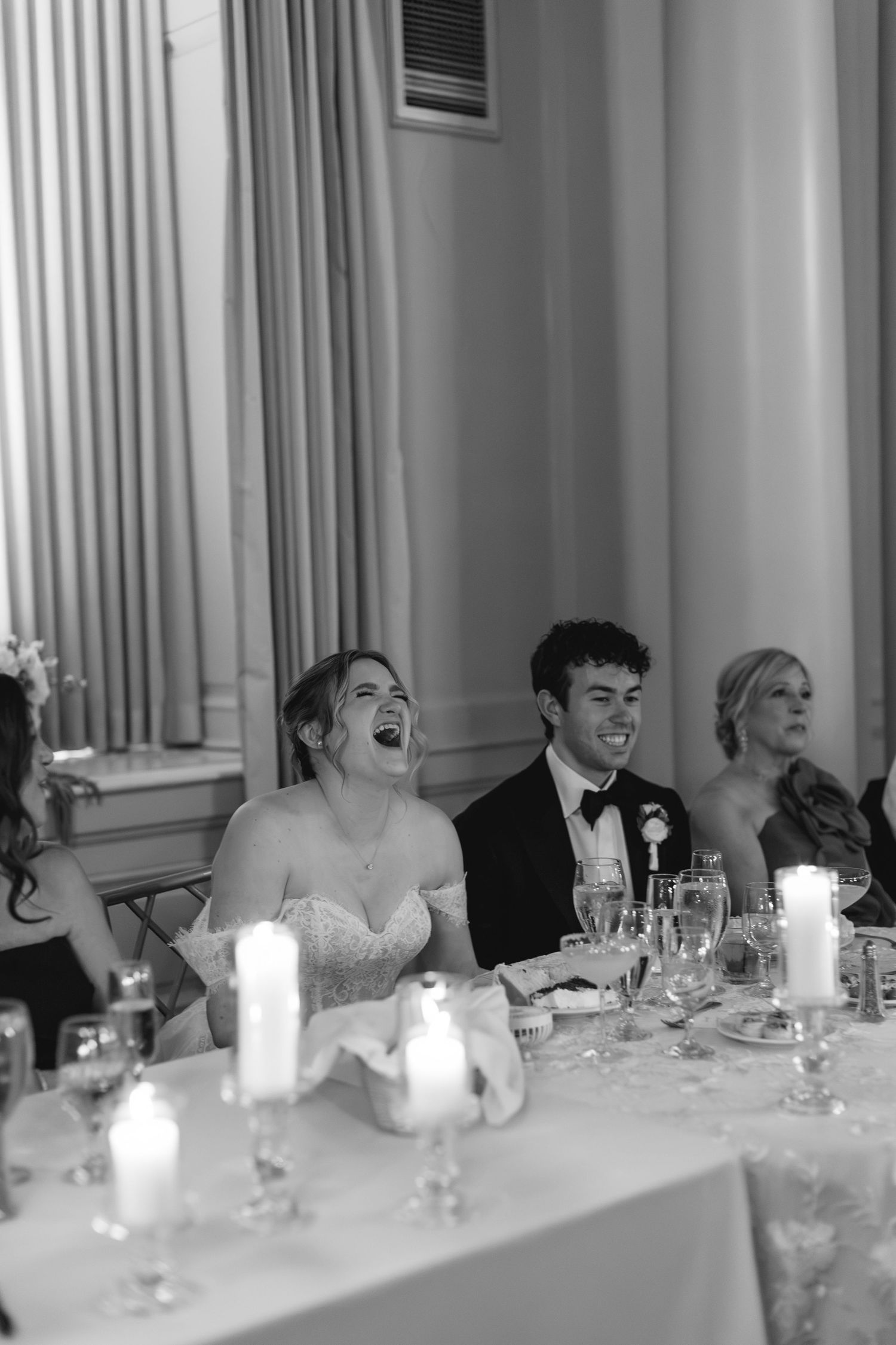 Black and white photo of a laughing bride and smiling groom seated at a candlelit wedding reception table.