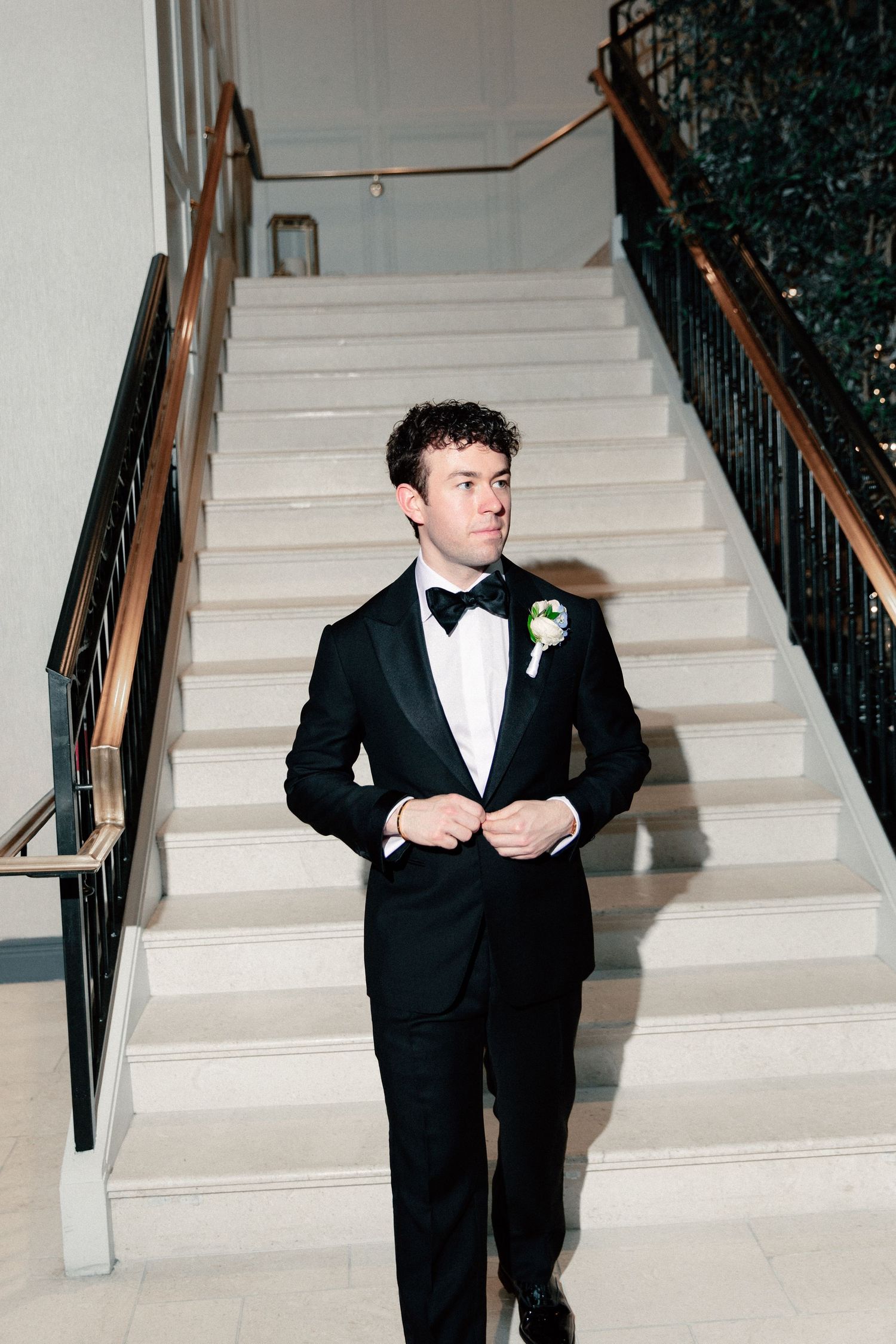 A dapper groom in a black tuxedo with boutonniere buttons his jacket while standing at the base of an elegant staircase.
