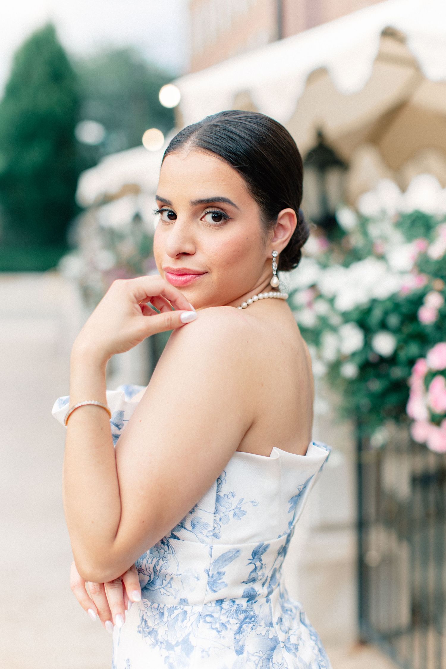 Elegant woman in a blue floral strapless dress with pearl necklace posing outdoors near flowers.