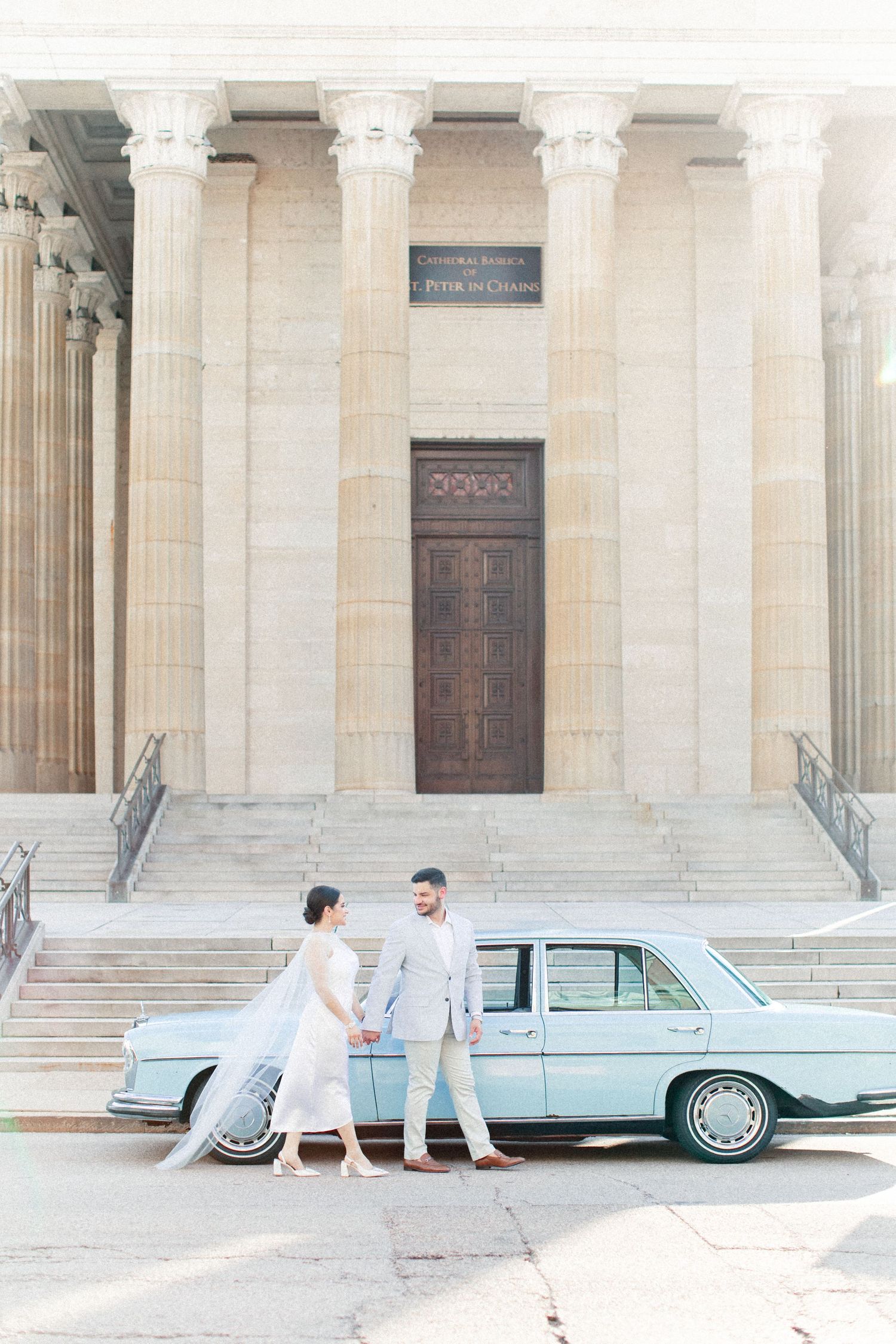 Bride and groom hold hands beside a vintage light blue car in front of a grand neoclassical church with tall columns.
