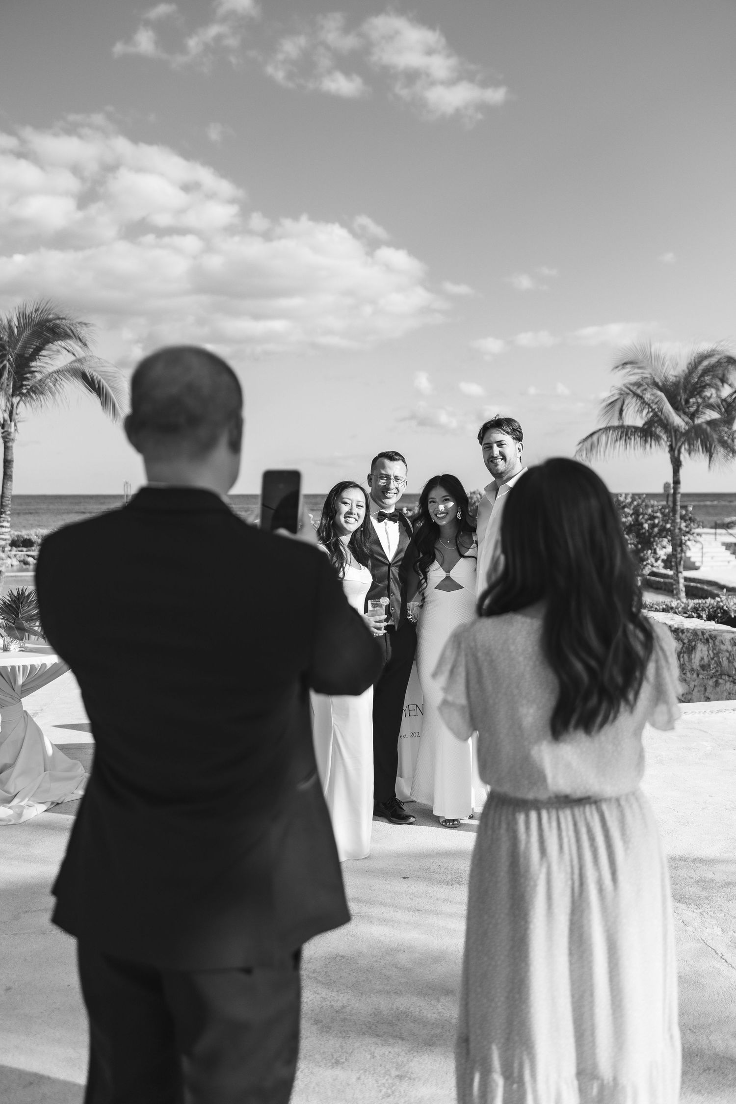 Black and white photo of wedding guests posing for a photo on a tropical resort terrace with palm trees and ocean views.